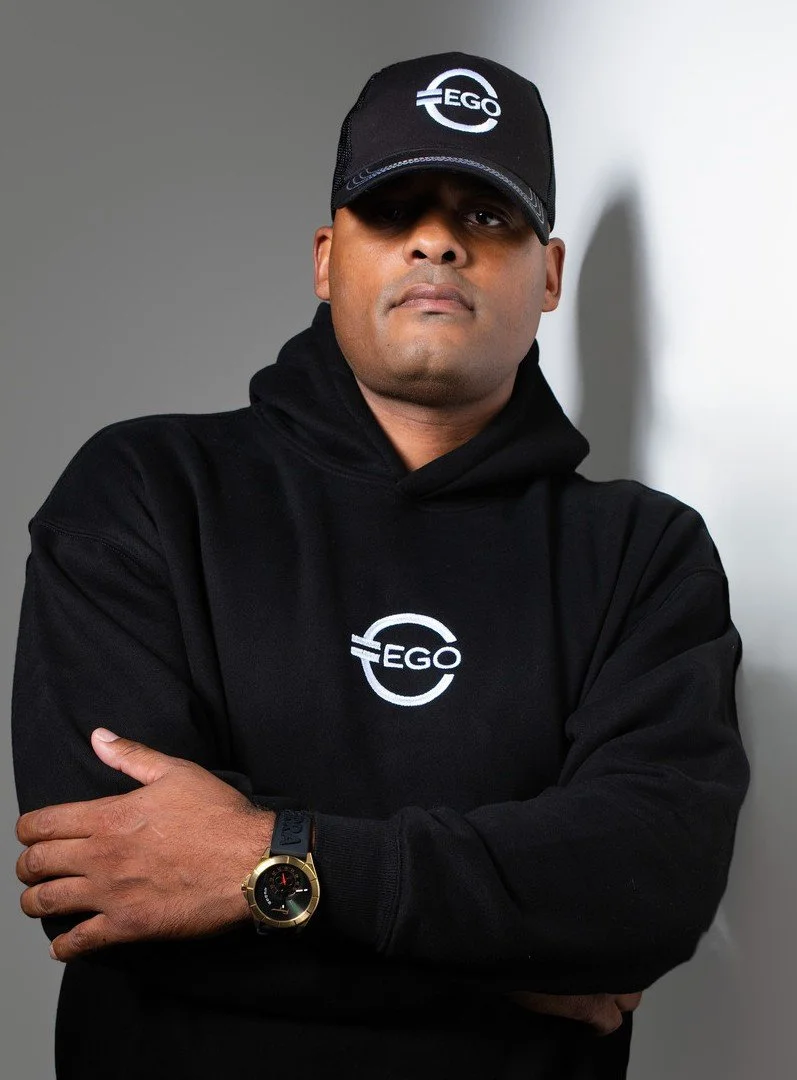 A man wearing a black cap and hoodie with the logo 'EGO' poses with arms crossed against a gray wall.