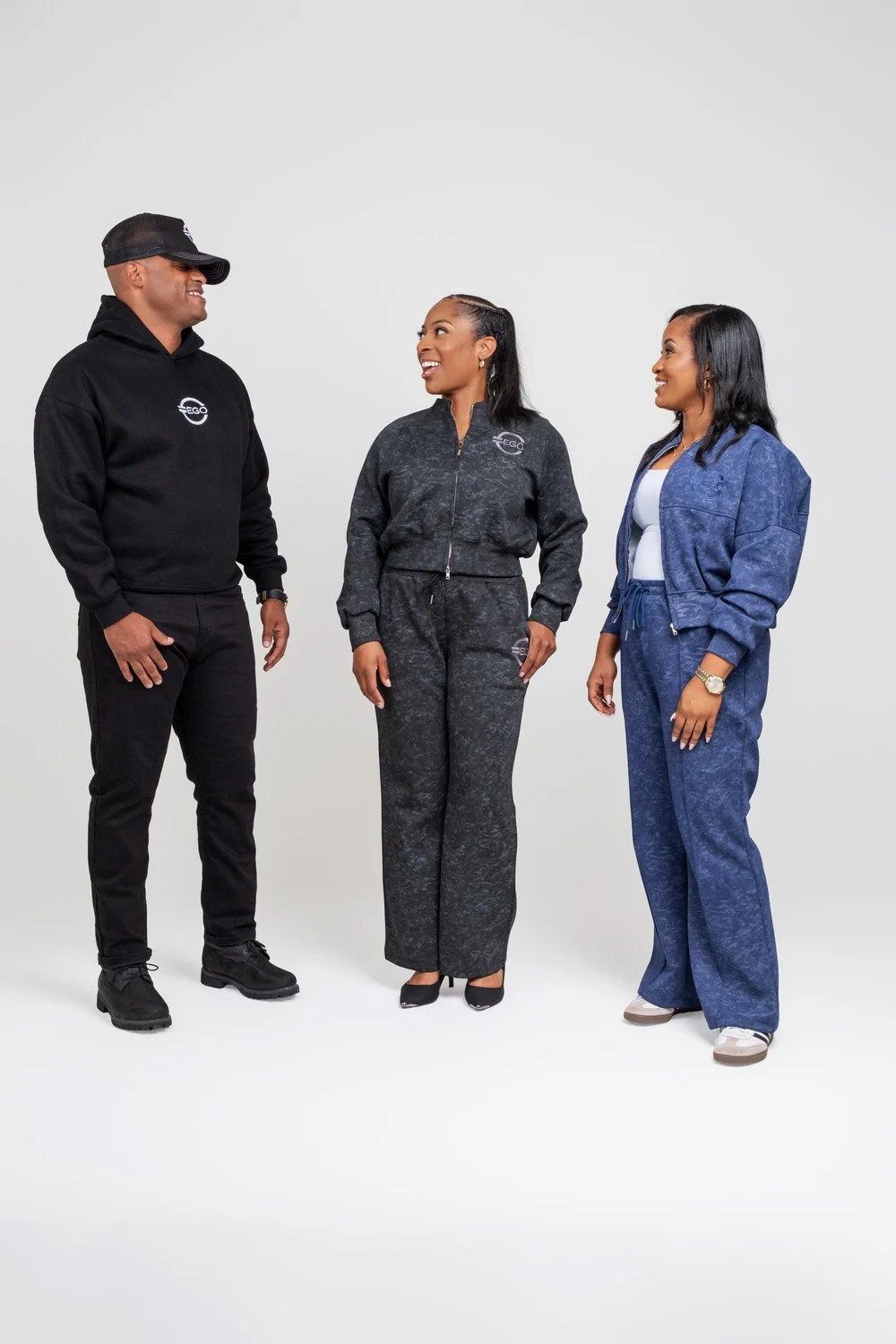 Three people standing and smiling at each other, dressed in casual and sporty clothing, against a plain white background.
