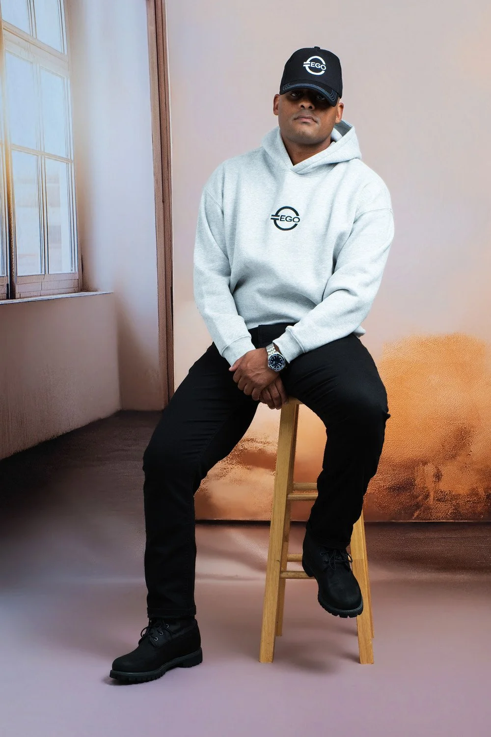 A man wearing a black cap, gray hoodie, black pants, and black boots sitting on a wooden chair in front of a backdrop with a cloud and sky scene, next to a window with sunlight.