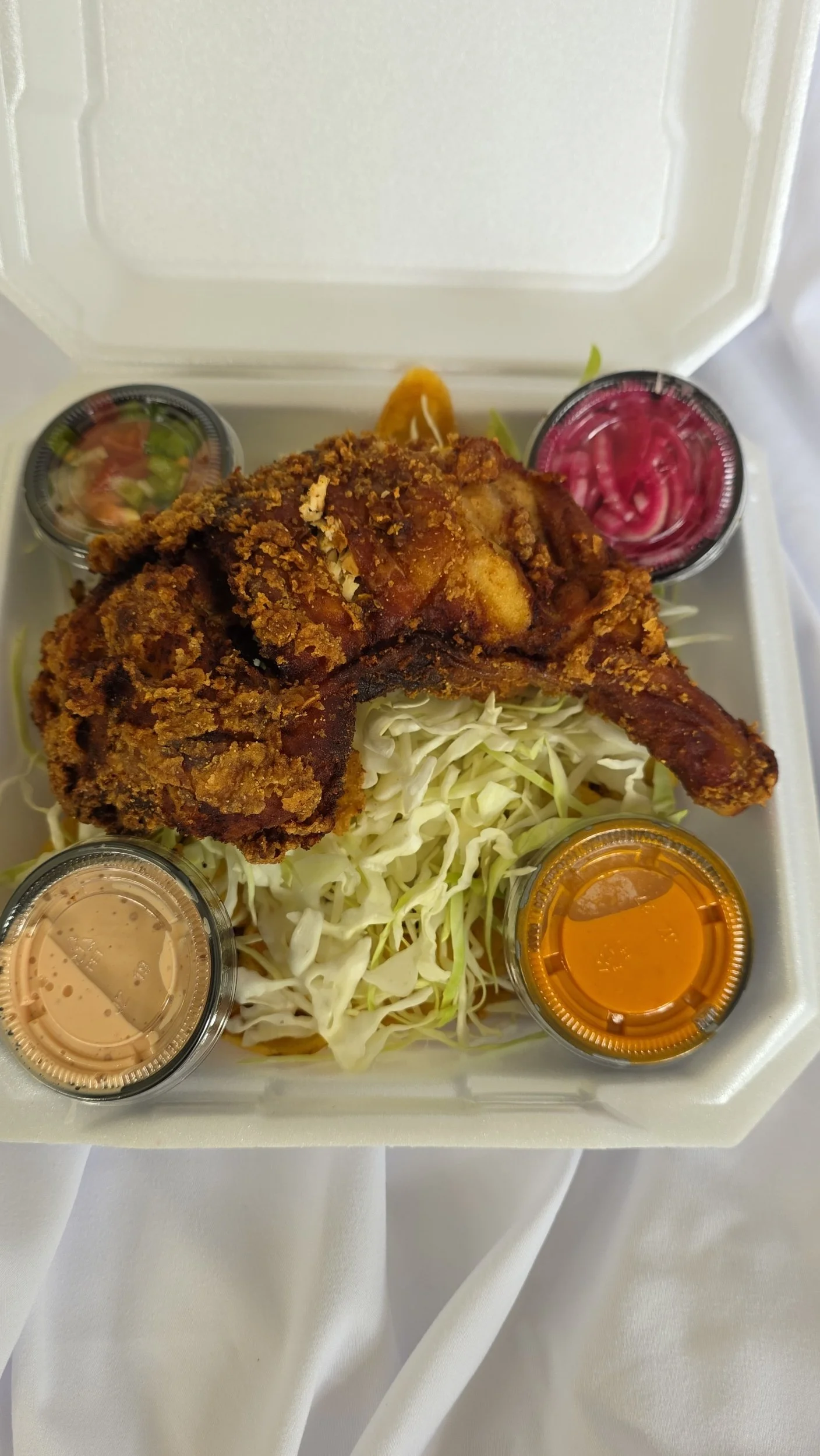 Fried chicken leg on shredded cabbage, served with small containers of sauce, pickles, and dressing in foam container.