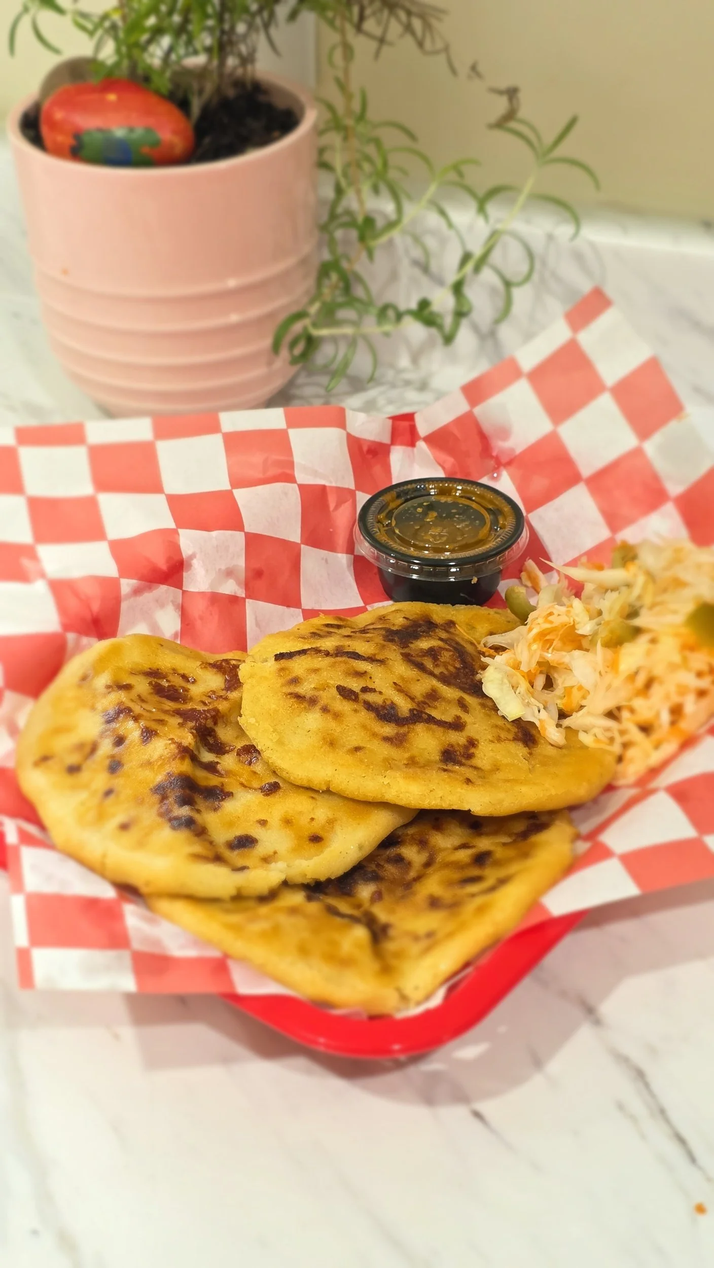 Cheese quesadilla with side of coleslaw, served on red-checkered paper in a red basket, with a container of sauce, on a white marble table.