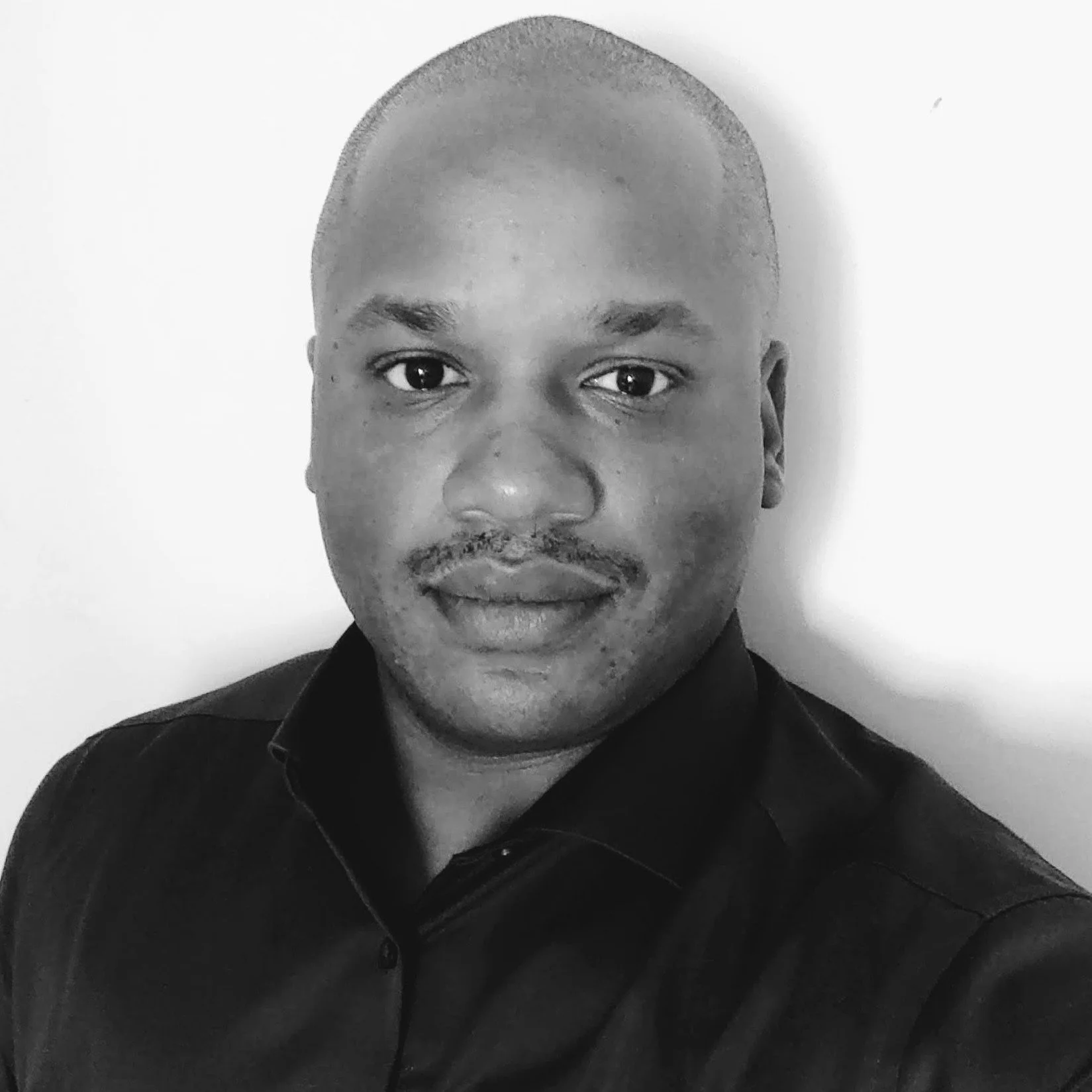 A black-and-white close-up portrait of a man with short hair and facial hair, wearing a dark shirt, standing against a light background.