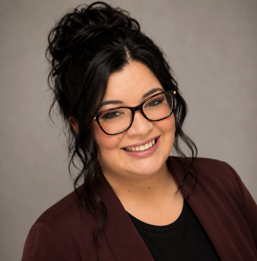 A woman with long dark hair in a messy bun, wearing glasses, a black shirt, and a maroon blazer, smiling against a neutral background.