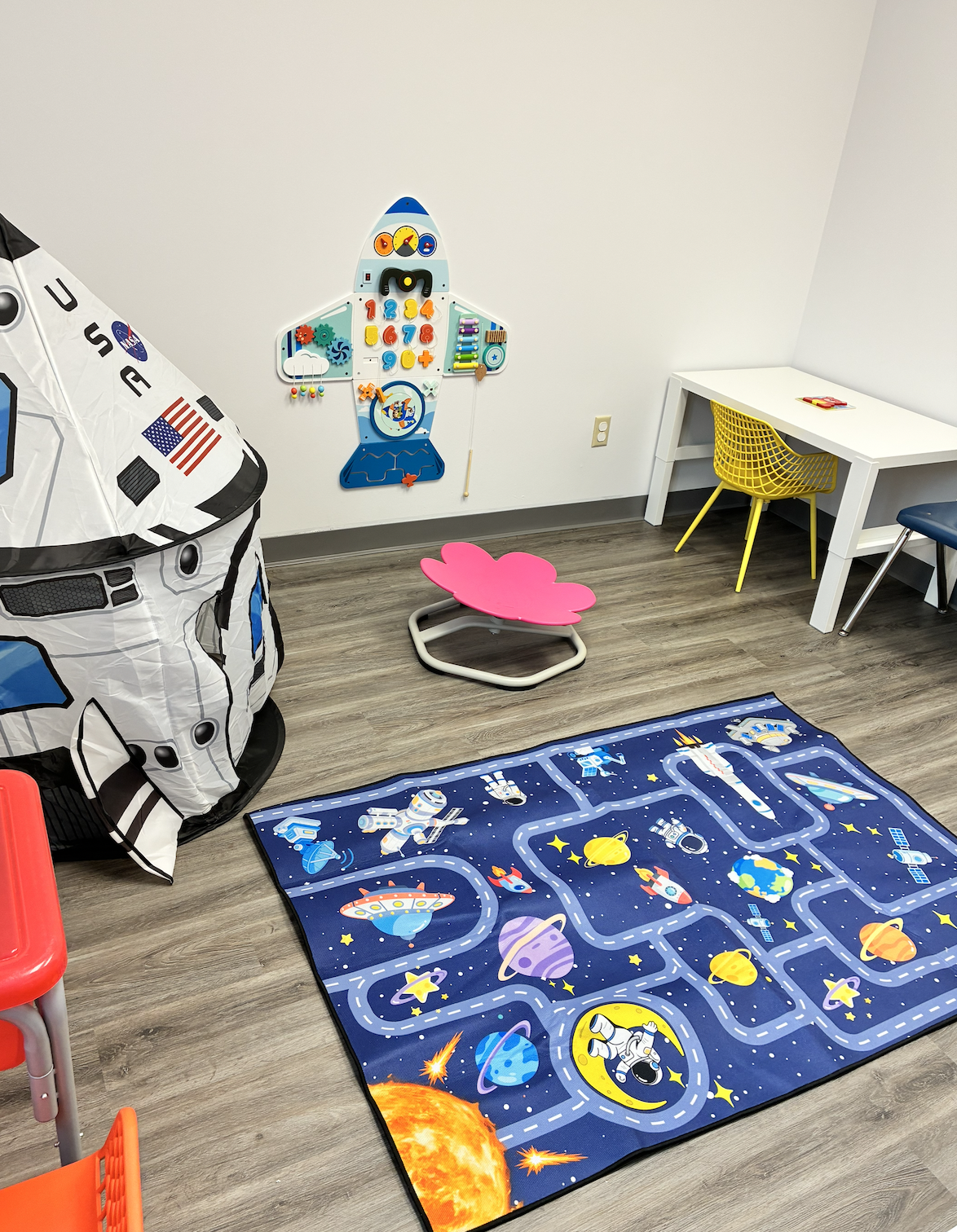 Children's playroom with a space-themed rug, star and planet illustrations, a pink footstool shaped like a flower, a white desk with a yellow chair, a fire truck tent, and space-themed wall decoration.