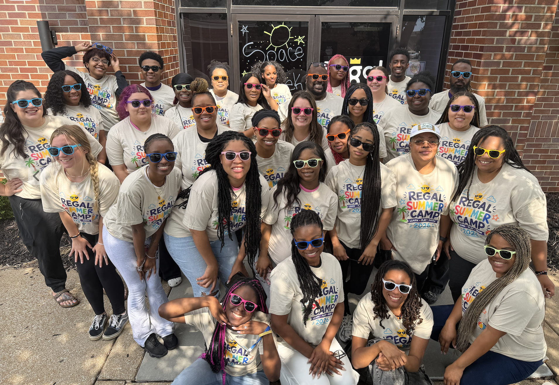 Group of diverse young people wearing sunglasses and matching t-shirts that say 'Regal Summer Camp 2026,' standing outside in front of a brick building with glass doors that have space-themed artwork and writing. Some are smiling and posing for the photo.