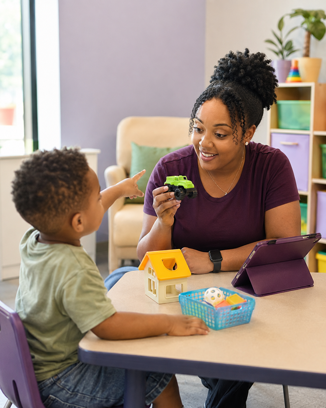 A woman and a young boy sitting at a table in a room with colorful shelves, playing with toys. The woman is holding a green toy truck and smiling at the boy, who is pointing at her. There is a small house model, a basket with toys, and a tablet on the table.