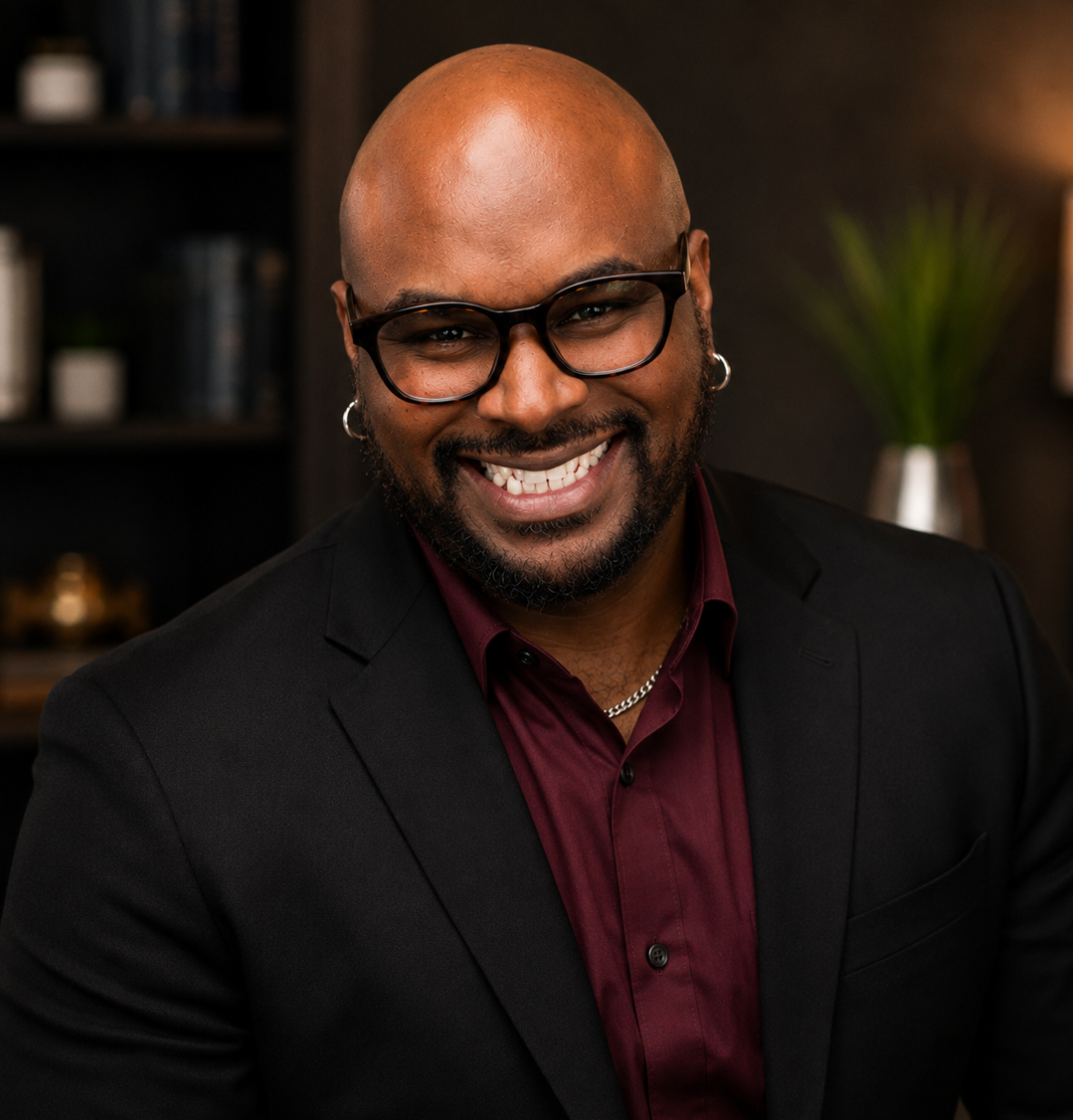 A smiling man with glasses, earrings, and a beard, dressed in a black suit and maroon shirt, in a modern indoor setting with a dark background, bookshelves, and a potted plant.