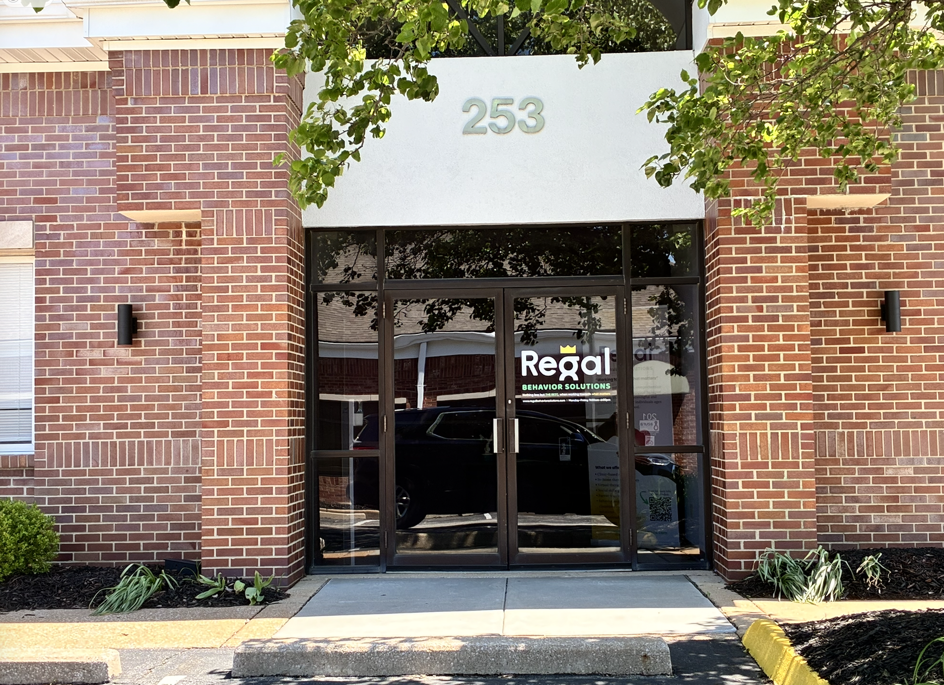 The brick storefront of a building with the number 253 displayed above the entrance. The entrance has glass doors with a 'Regal Behavior Solutions' sign. There are trees, plants, and a parking lot in front.