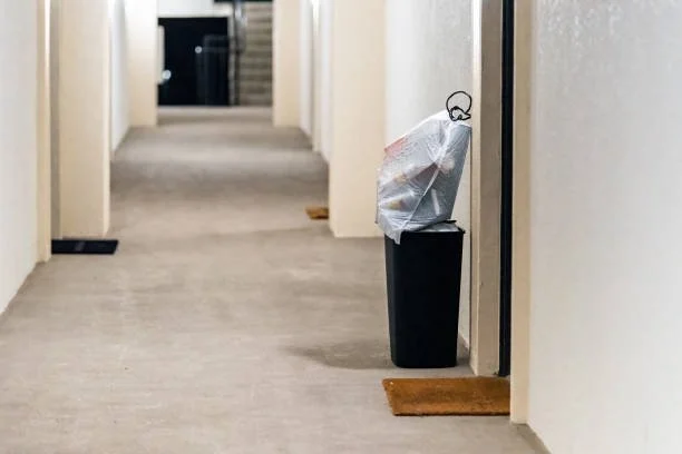 A hallway with beige walls and gray carpet, with a trash can outside a door that has a plastic bag with trash and a wire handle on top. There is a doormat in front of the door.