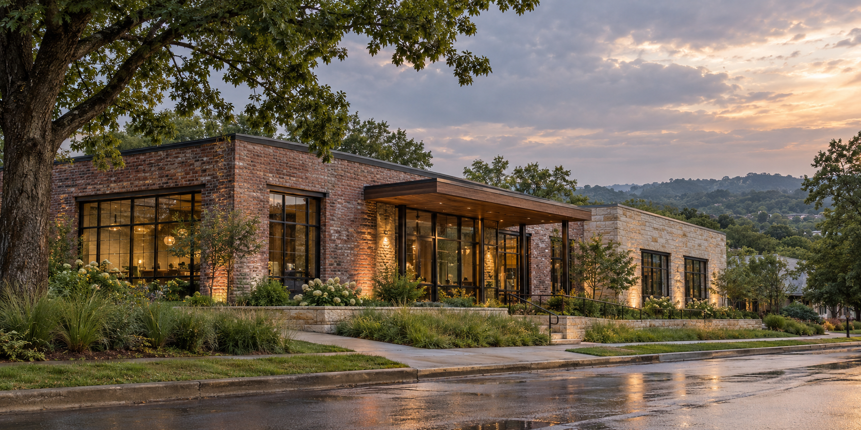 A modern brick and stone building at sunset, with large glass windows, surrounded by greenery and trees, and wet pavement in the foreground.