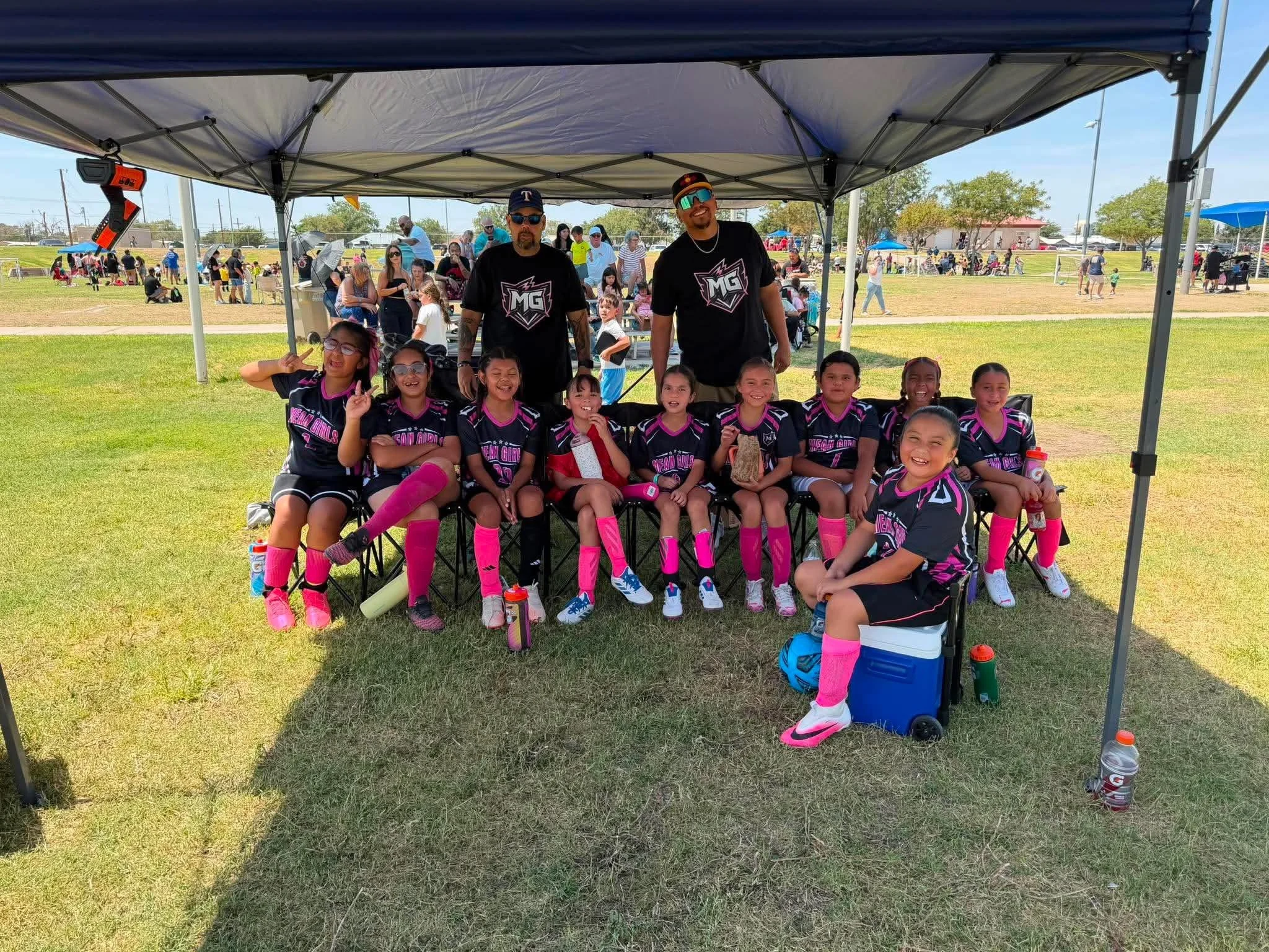 A group of young girls in black and pink sports uniforms sitting under a canopy with two adult men standing behind them at a sports event in a park on a sunny day. Many spectators are visible in the background.