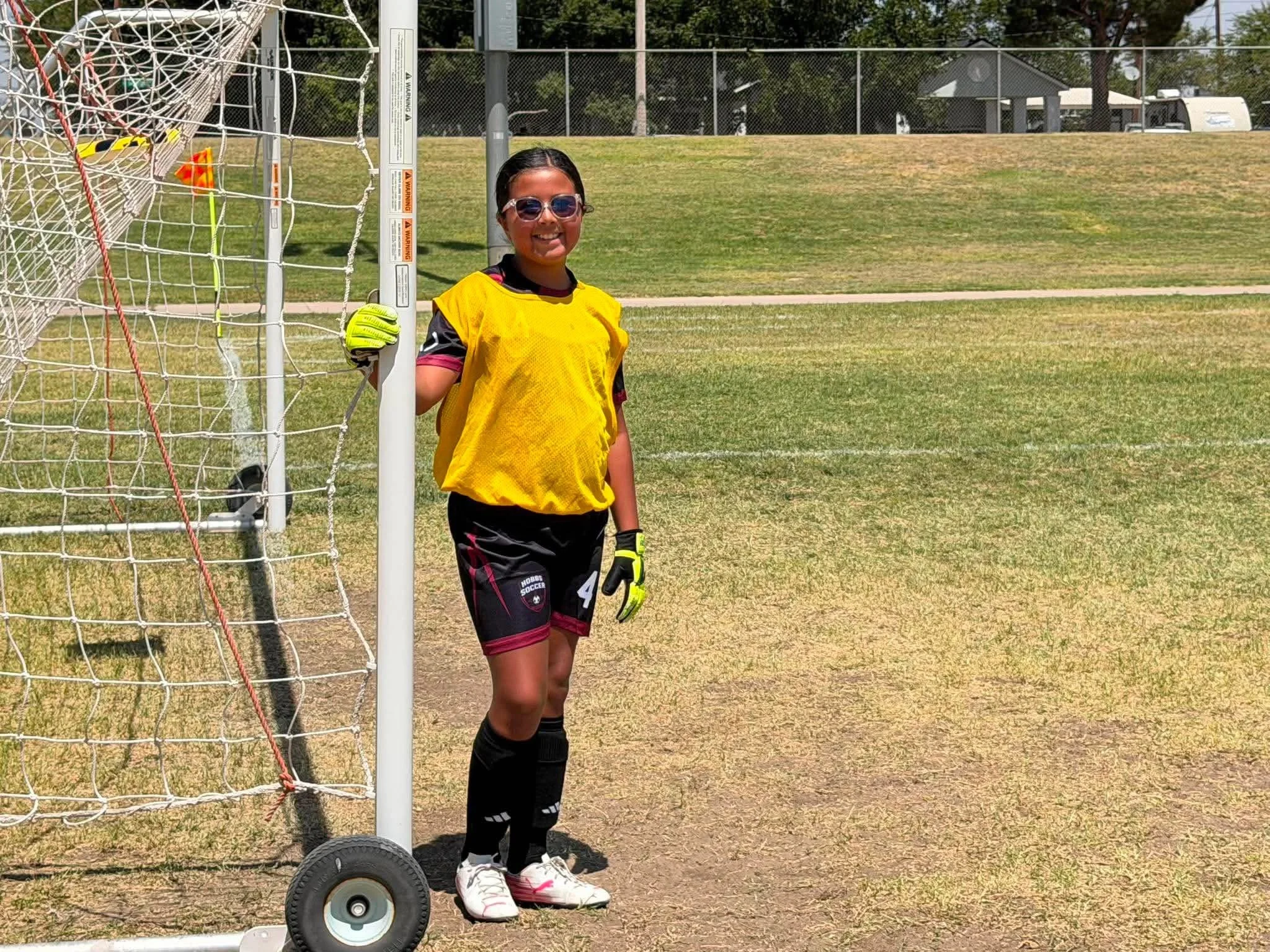 Young girl in soccer uniform and sunglasses standing next to soccer goal, holding goalpost on sunny day at park.