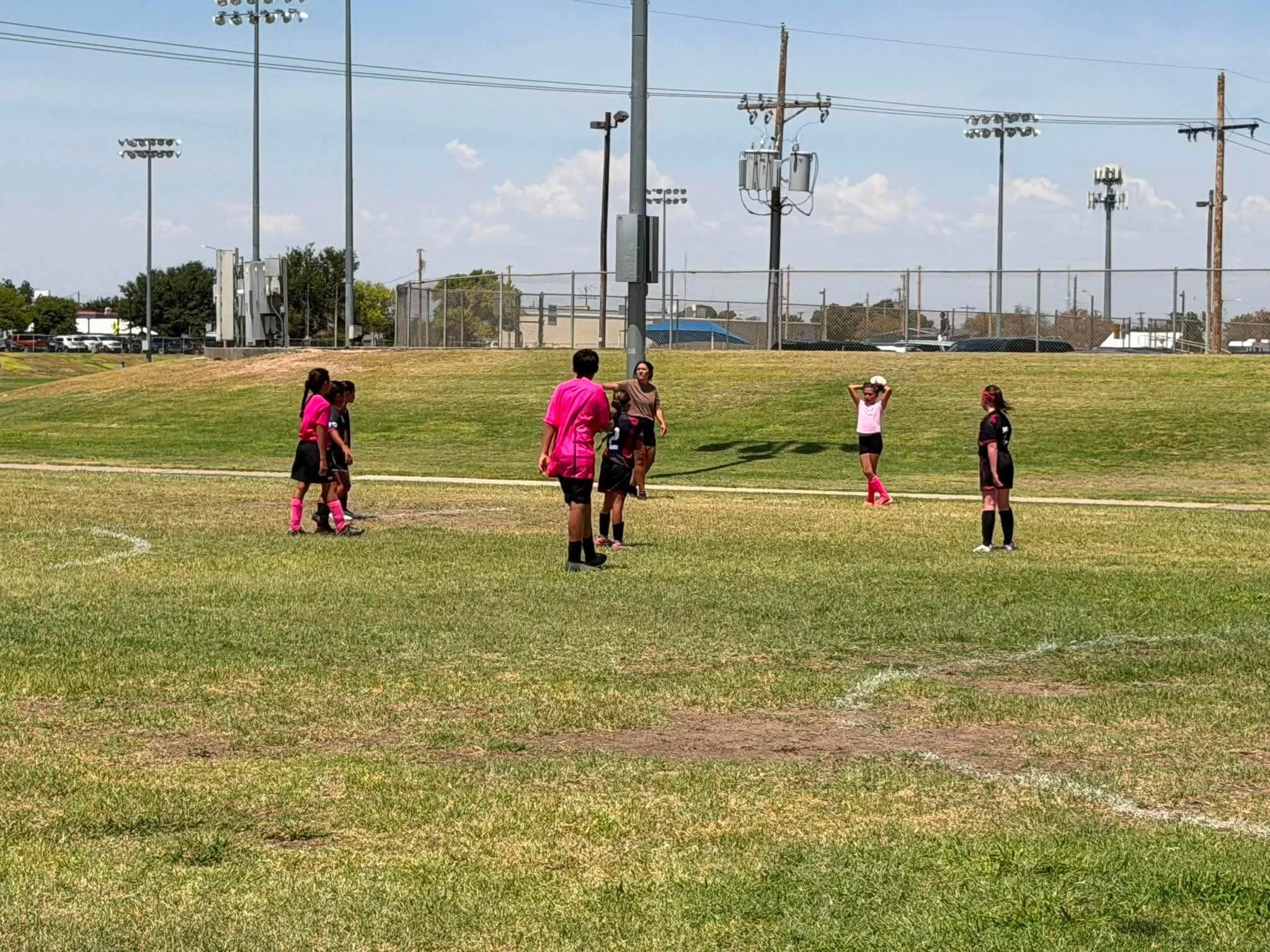 A group of young girls and women standing on a grassy field, some wearing sports uniforms, with a chain-link fence, utility poles, and light poles in the background under a blue sky with scattered clouds.