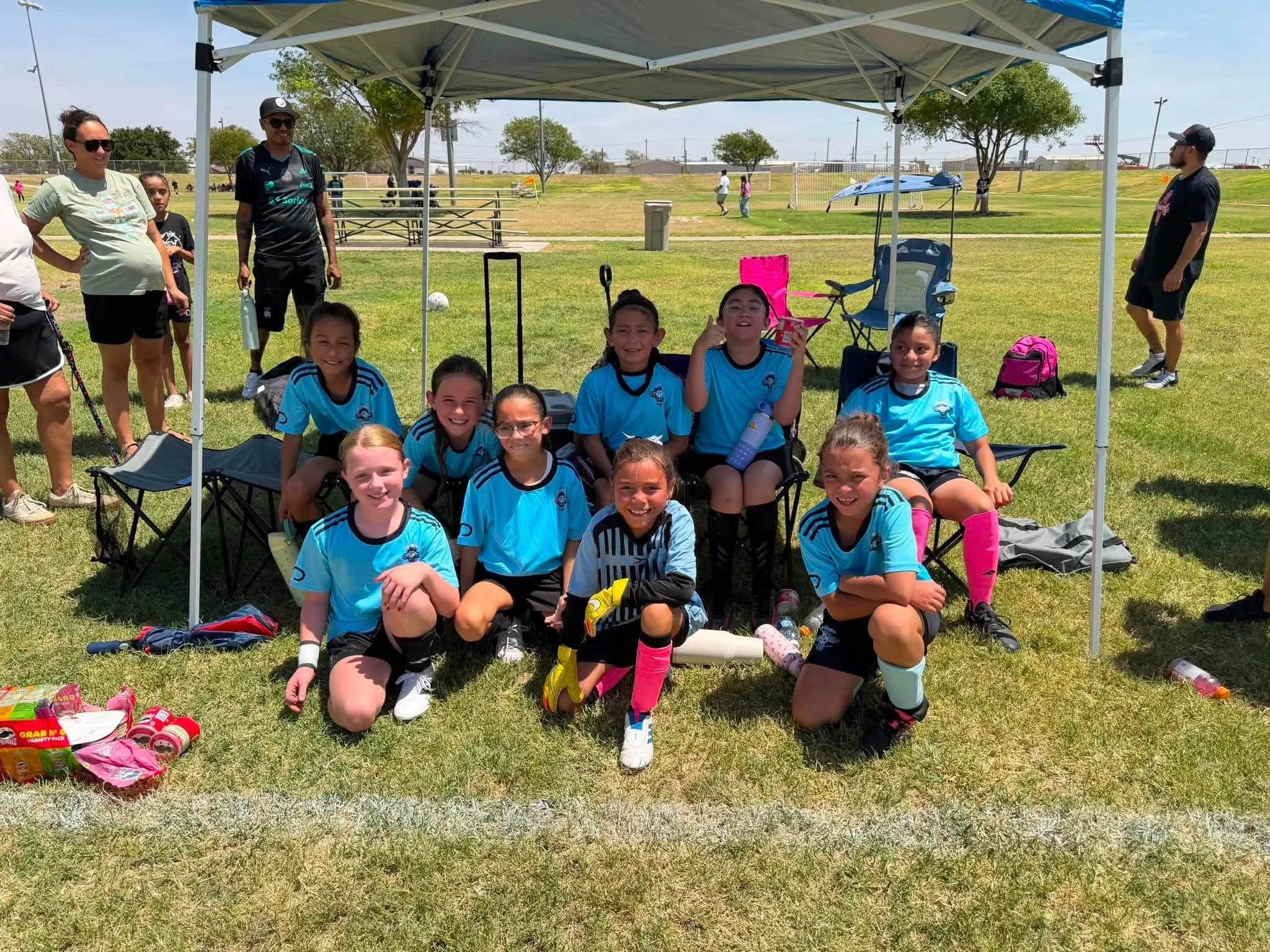 Young girls in blue soccer jerseys sitting and smiling in front of a shaded canopy at a soccer field, with more people and children playing in the background.