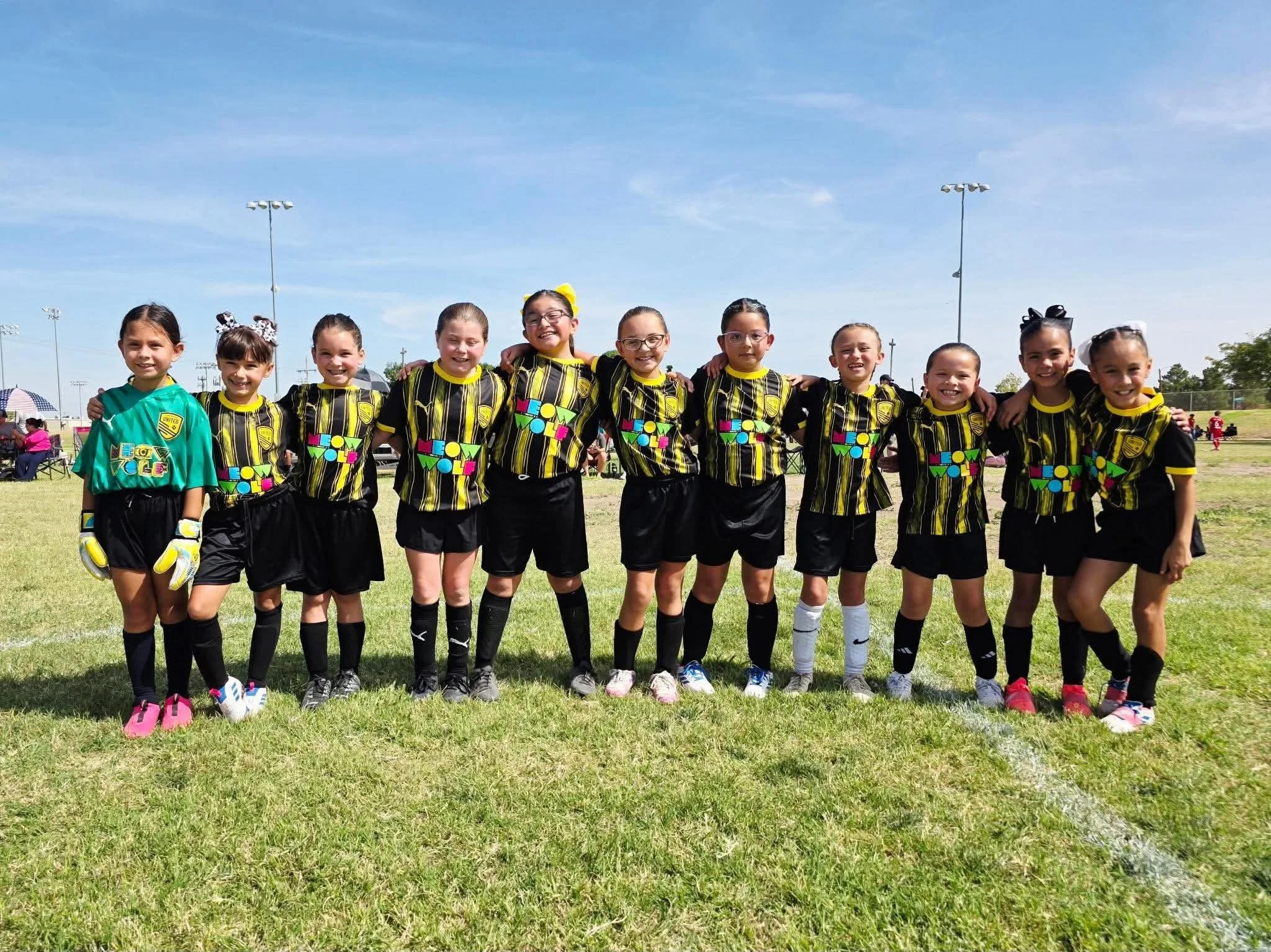 A group of ten young girls standing on a grassy field, dressed in soccer uniforms, with arms around each other, smiling for a photo during a daytime soccer event.