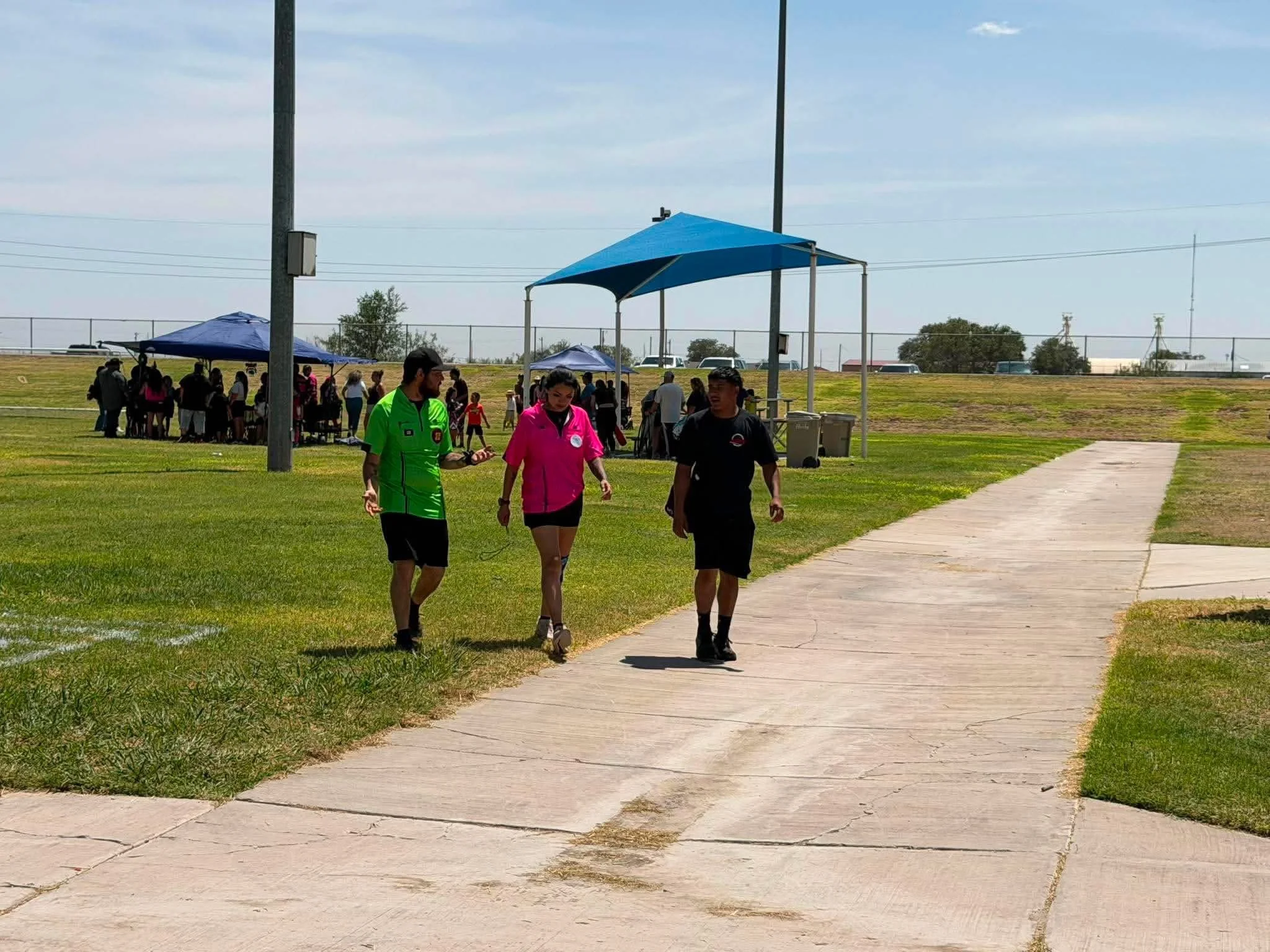 Several people walking outside near a blue canopy tent on a grassy field, with some standing and others walking on a concrete path under a partly cloudy sky.