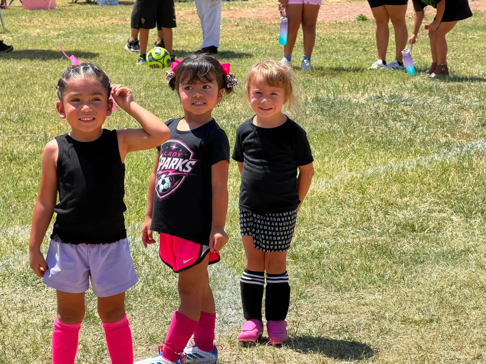 Three young girls standing on grass, wearing sports attire, with other children in the background playing with a soccer ball and water bottles on a sunny day.