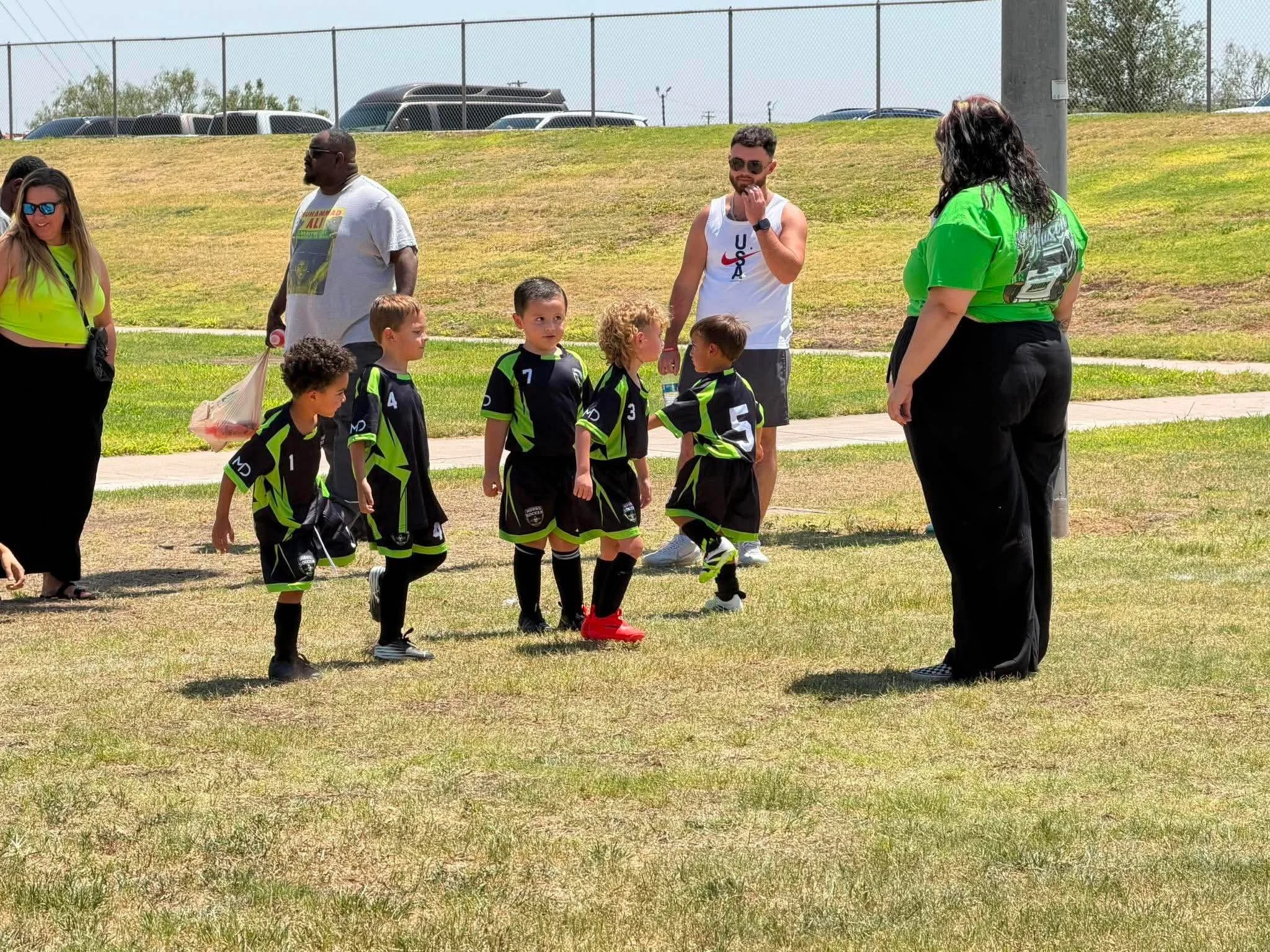 A group of children and adults are gathered outdoors on a grassy field. The children are in black and green sports uniforms, walking in a line. The adults are standing nearby, some with sunglasses. A fence and parked cars are visible in the backgroun