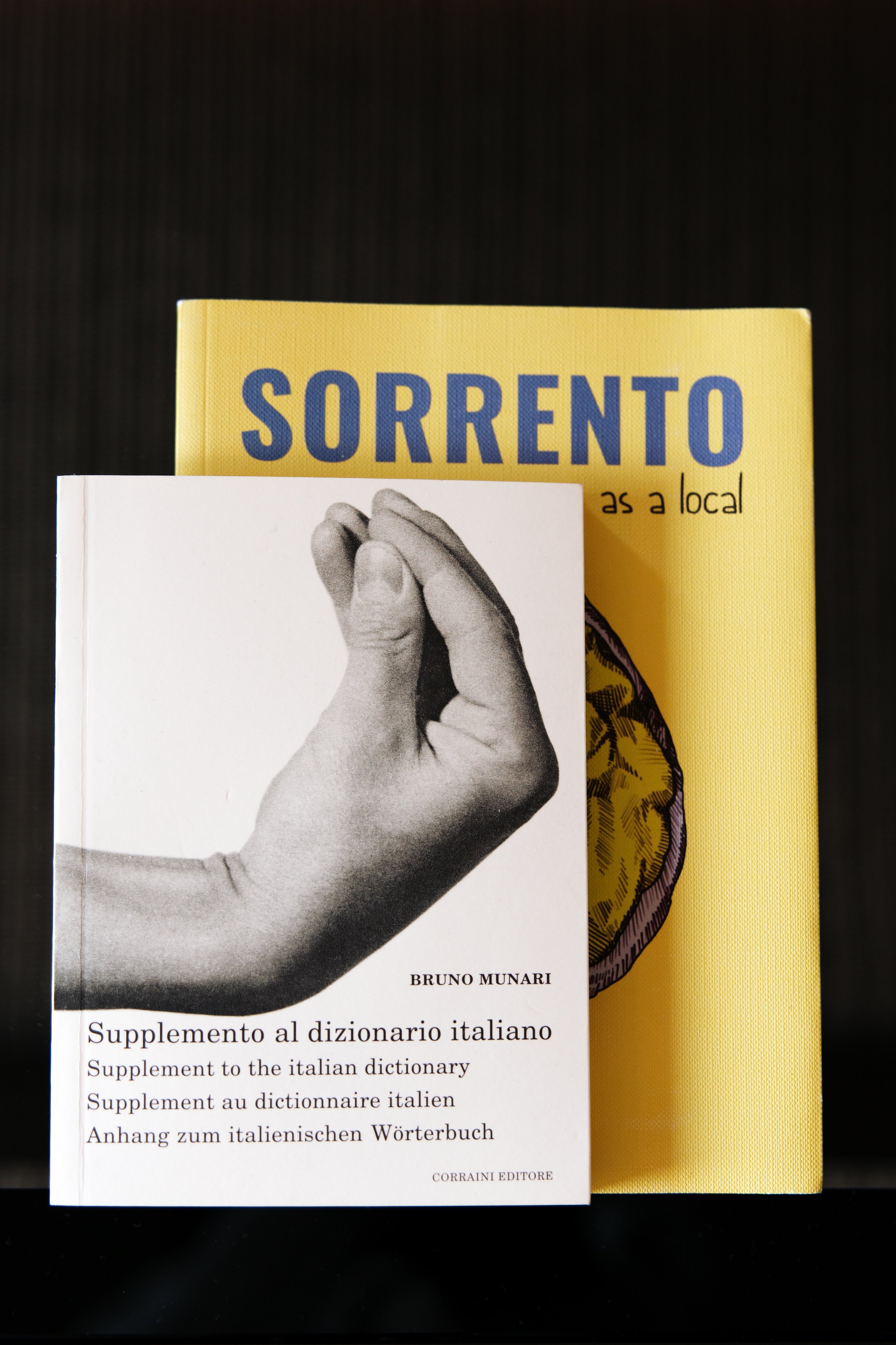 A black and white book cover featuring a hand holding a smaller hand, titled "Supplemento al dizionario italiano" by Bruno Munari. Behind the book, there is a yellow book titled "Sorrento as a local."