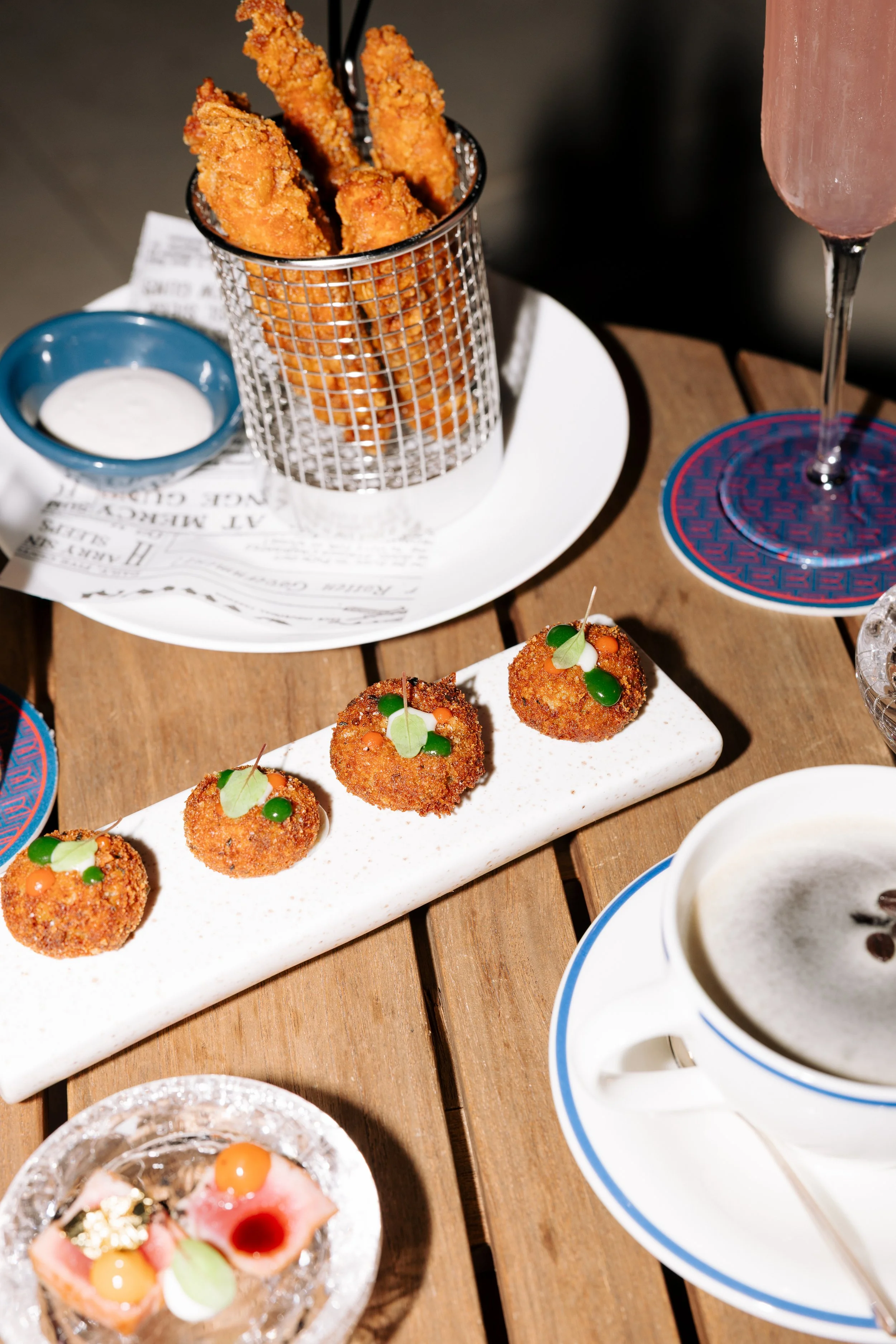 Fried chicken strips in a metal basket with a small cup of dipping sauce on a white plate, decorated appetizers with various toppings, pink drink in a tall glass on a coaster, cup of coffee or tea in a white cup, all arranged on a wooden table.