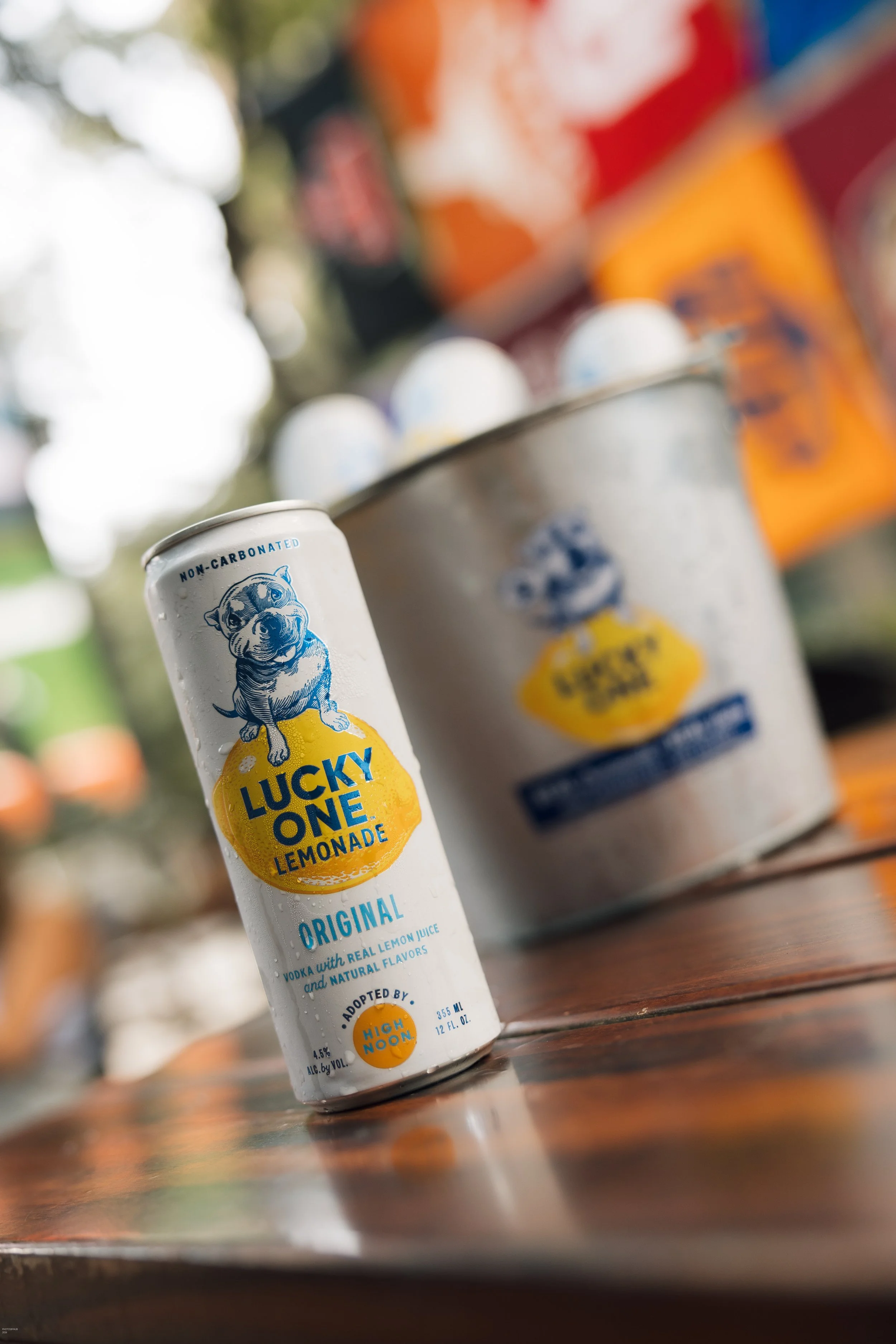 Close-up of a white can of Lucky One Lemonade with a blue bulldog logo, placed on a wooden surface. In the background, there are blurred cans and bottles, indicating a beverage display or cooler.