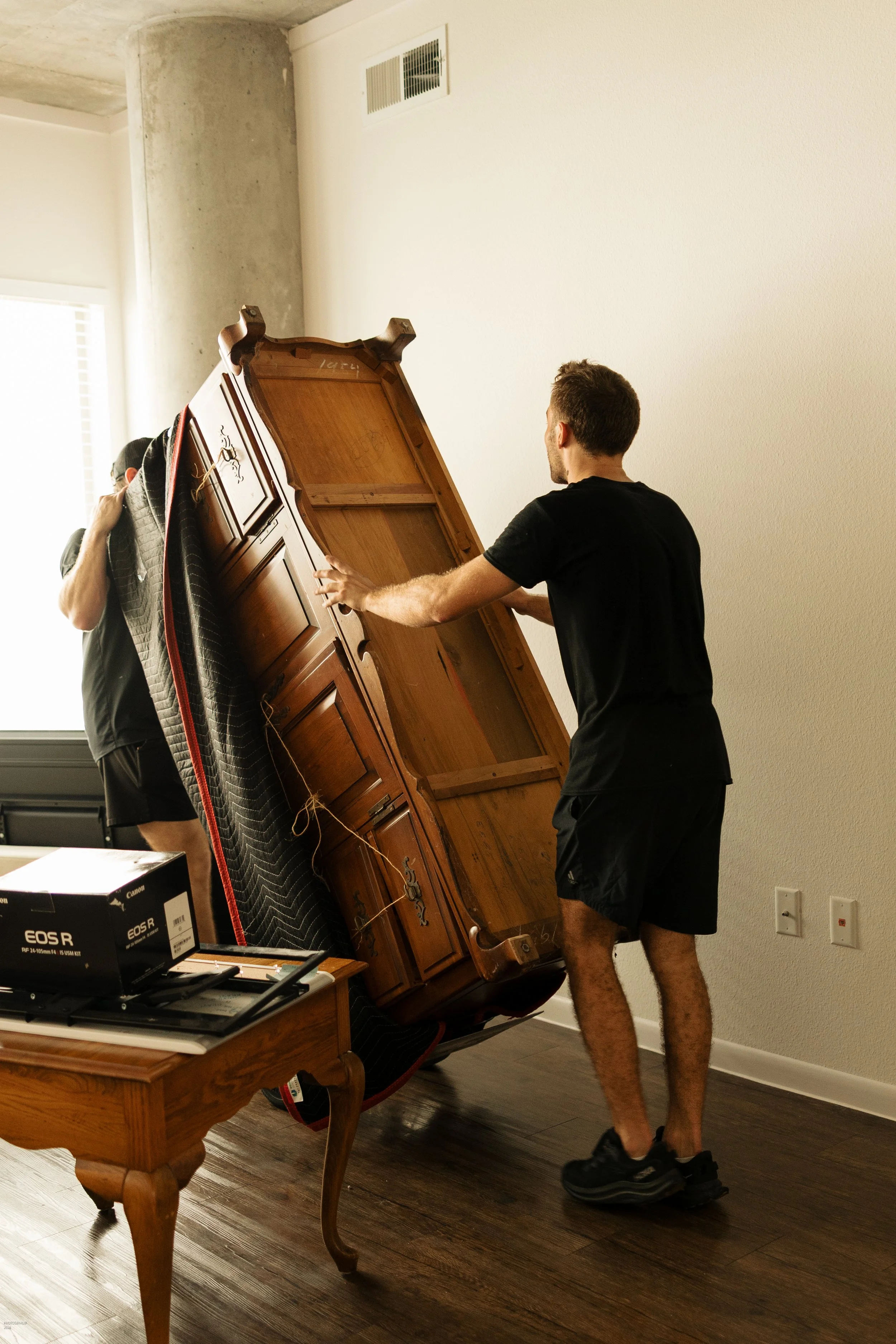 Two men are moving a large, upright wooden cabinet in a room. One man is holding the top of the cabinet, and the other is supporting the side. One man is wearing a black t-shirt and shorts, and the other man is wearing a cap and black t-shirt. There is a desk with a Canon EOS R camera box and other electronic items on it in the foreground, and the room has light-colored walls and a dark wooden floor.