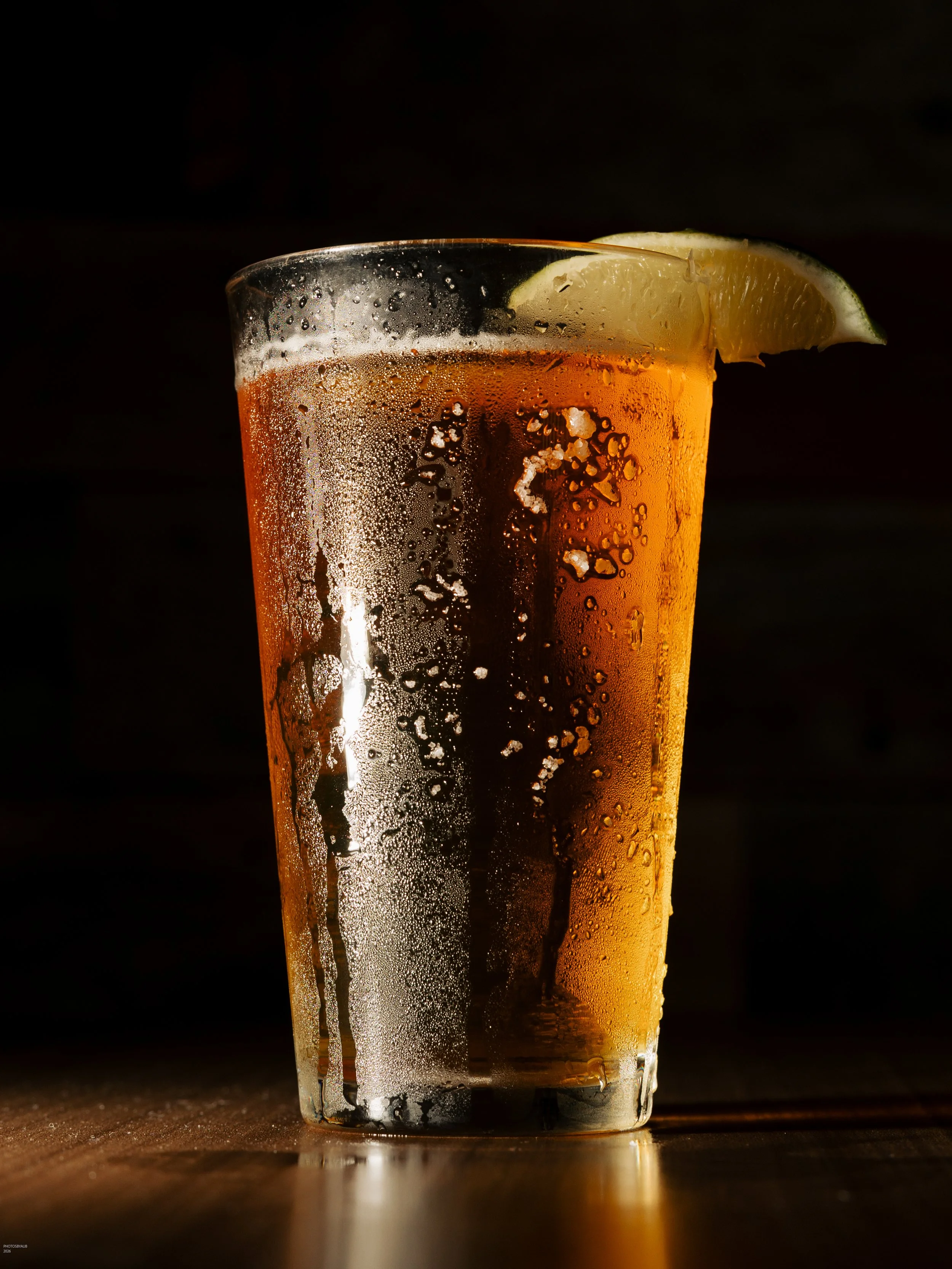 A glass of fizzy soda with ice and a lemon wedge on the rim, placed on a wooden surface against a dark background.