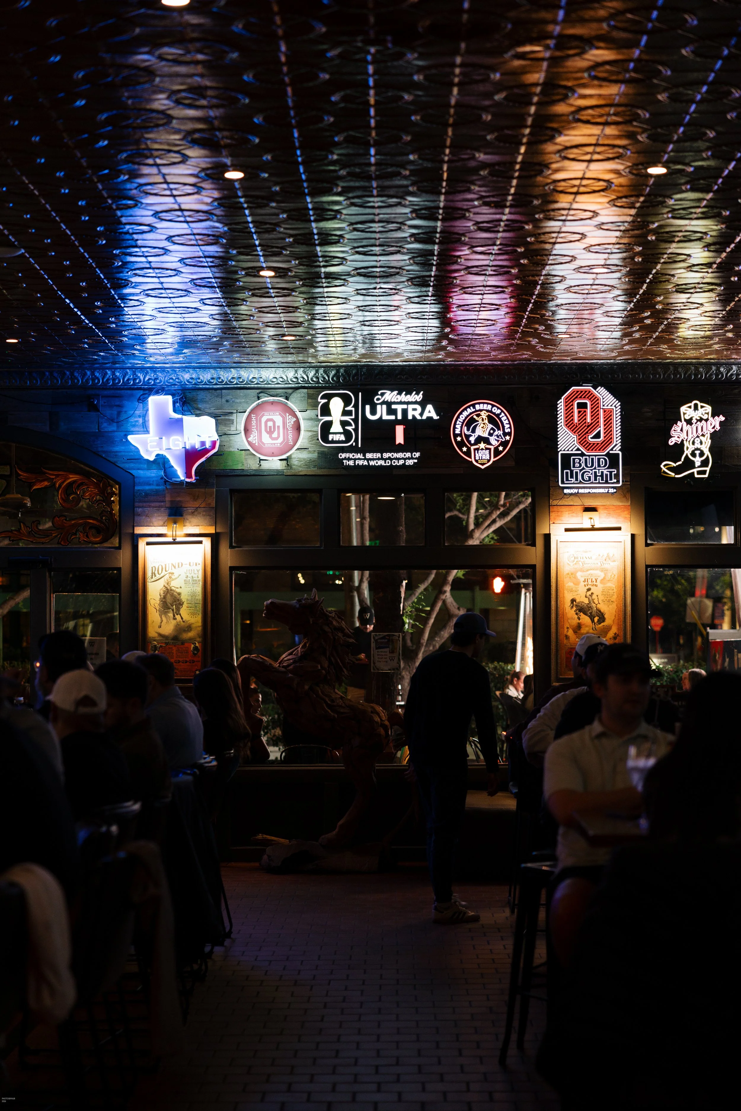 Night scene inside a bar or restaurant with neon signs on the wall, including the Texas state outline, Bud Light, and Oklahoma University logos. Indoor lighting reflects on the ceiling. People are sitting and standing, some in conversation. A statue or sculpture of a person holding a animal or creature is visible near the center.