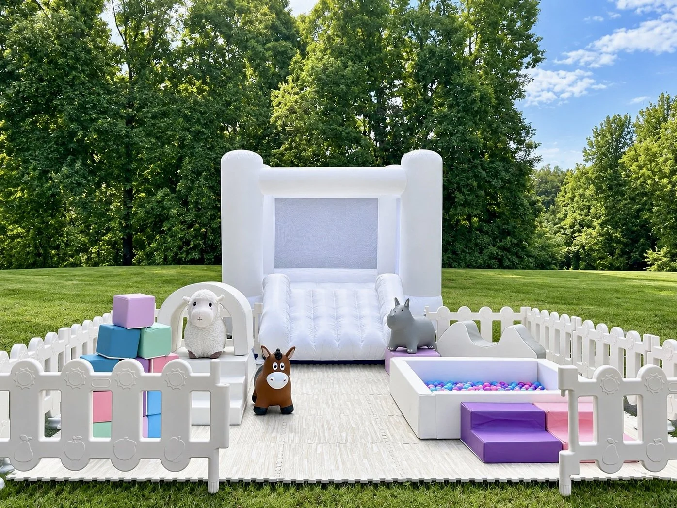 Outdoor inflatable bounce house with a slide, surrounded by a white picket fence, lawn, trees, and blue sky, decorated with toy animals and blocks.
