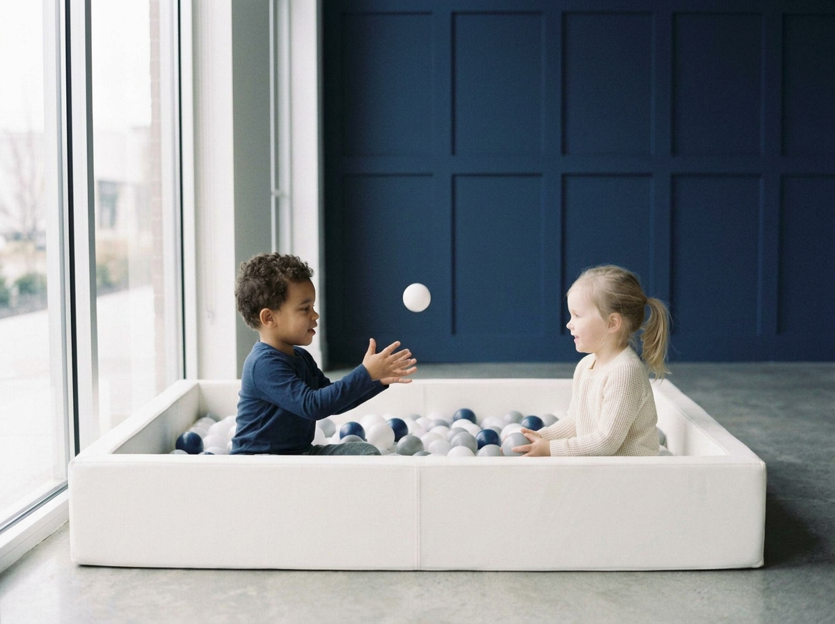 Two young children, a boy and a girl, playing together in a white ball pit near a large window, with the boy catching a ball and smiling at the girl.