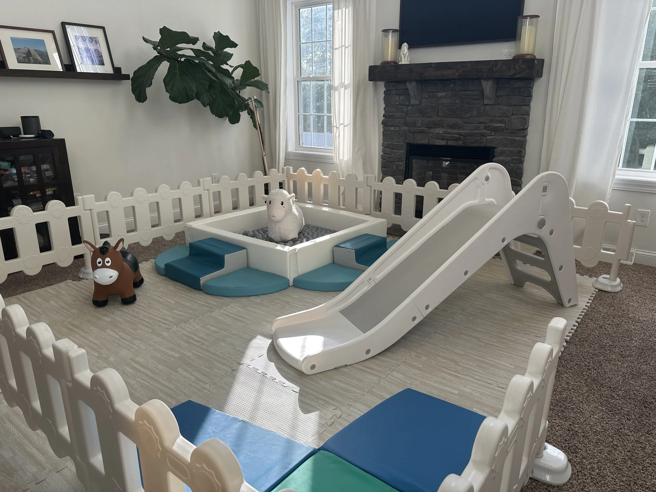 A children's play area with a white plastic slide, a white fence enclosure, and some stuffed toys including a sheep and a horse. There is a brick fireplace with a wooden mantel and a flat-screen TV above it. Large windows with white curtains let in natural light, and there is a large leafy plant in the corner.