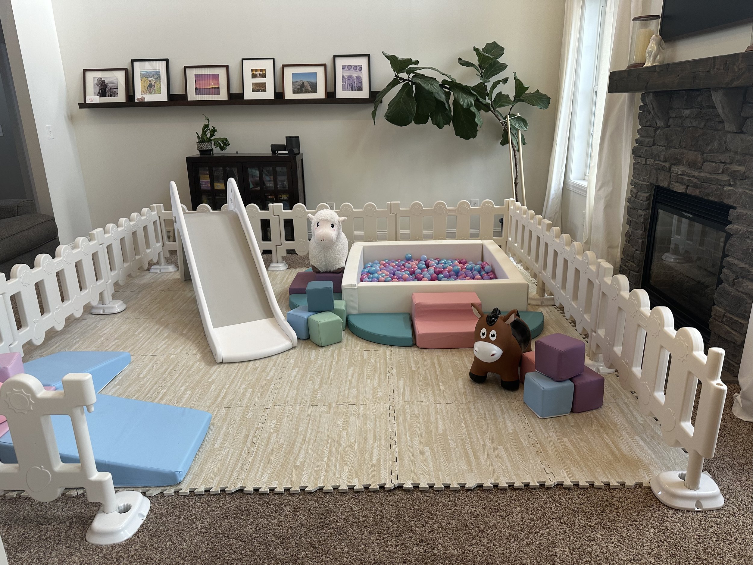 Play area with plastic fencing, a small slide, and toys including a plush llama, colorful blocks, and a toy horse in a living room with framed photographs on the wall, a large plant, a stone fireplace, and windows with curtains.