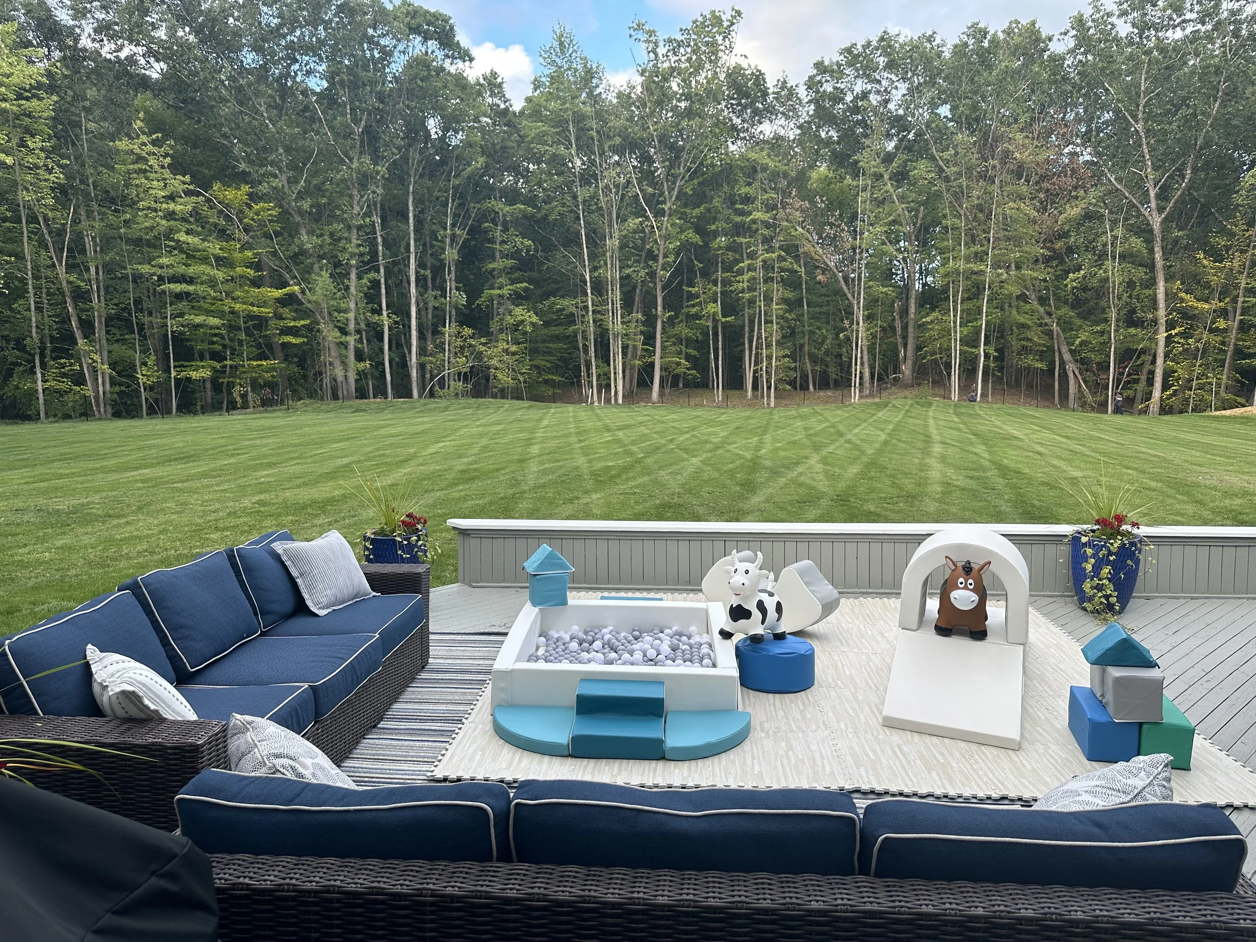 A backyard patio with blue outdoor furniture, children’s toys including a ball pit, cow and horse figures, and several potted plants. Beyond the patio is a large, well-maintained green lawn bordered by a line of trees and a blue sky with some clouds.