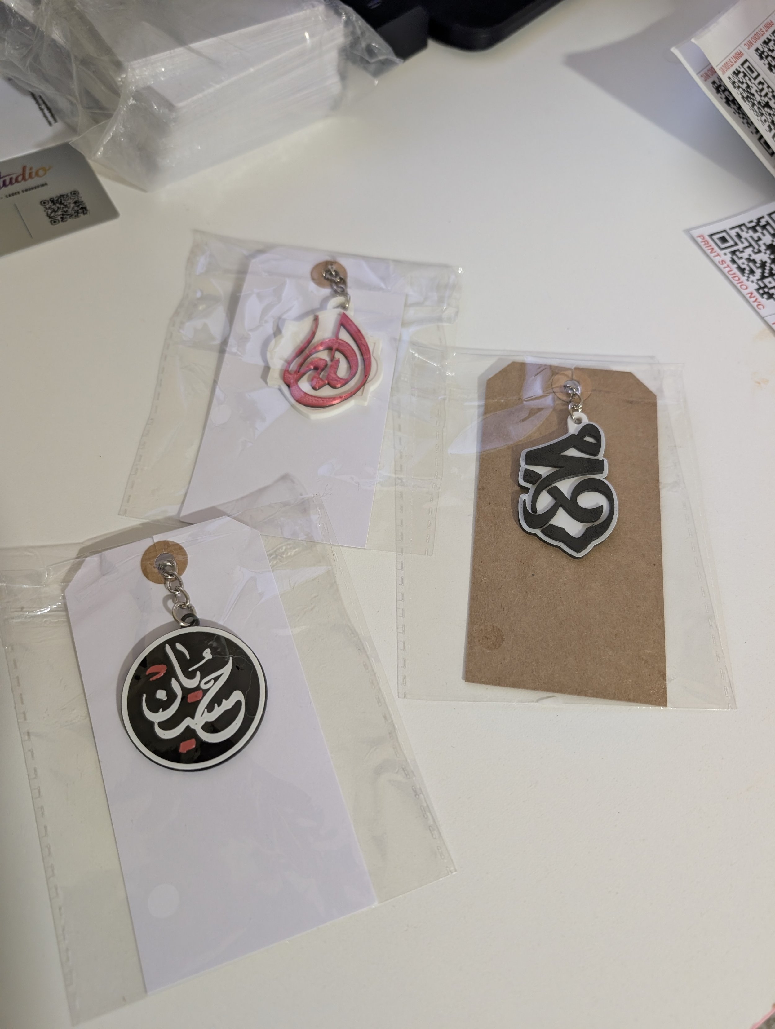 Two pairs of earrings in clear plastic bags, one with black and white circular design with red accents, the other with pink and white design, placed on a white surface with QR codes and packaging materials around.
