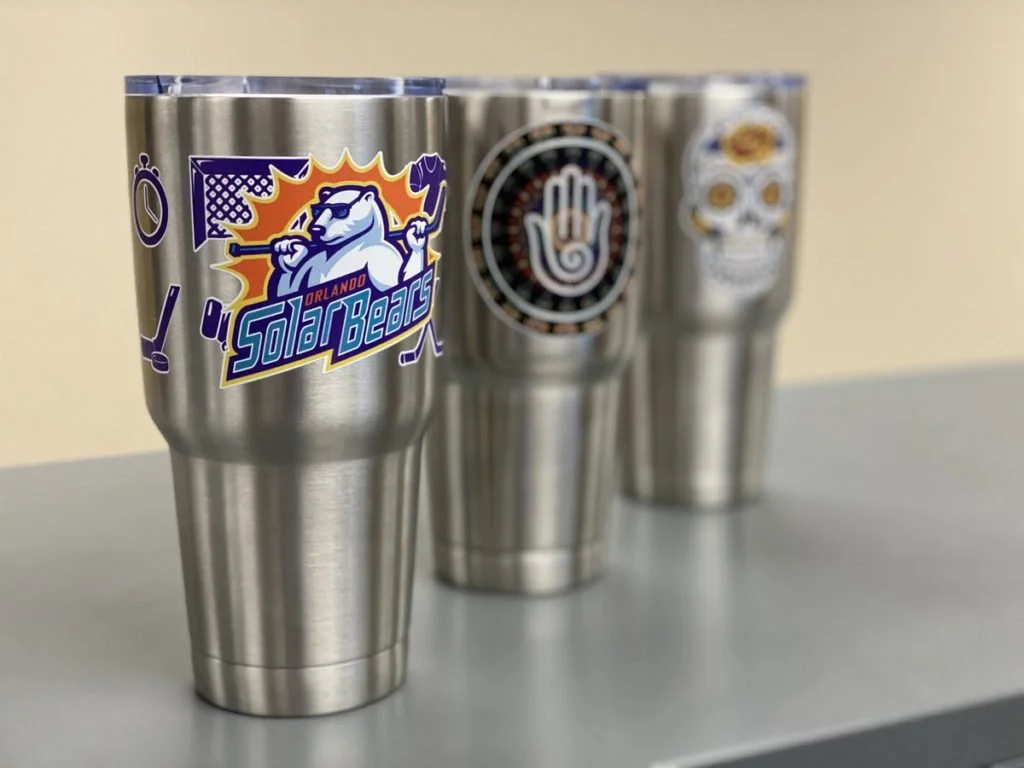 Three stainless steel tumblers with logos on them, one for the Orlando Solar Bears hockey team, one with a hand symbol, and one with a sugar skull design.