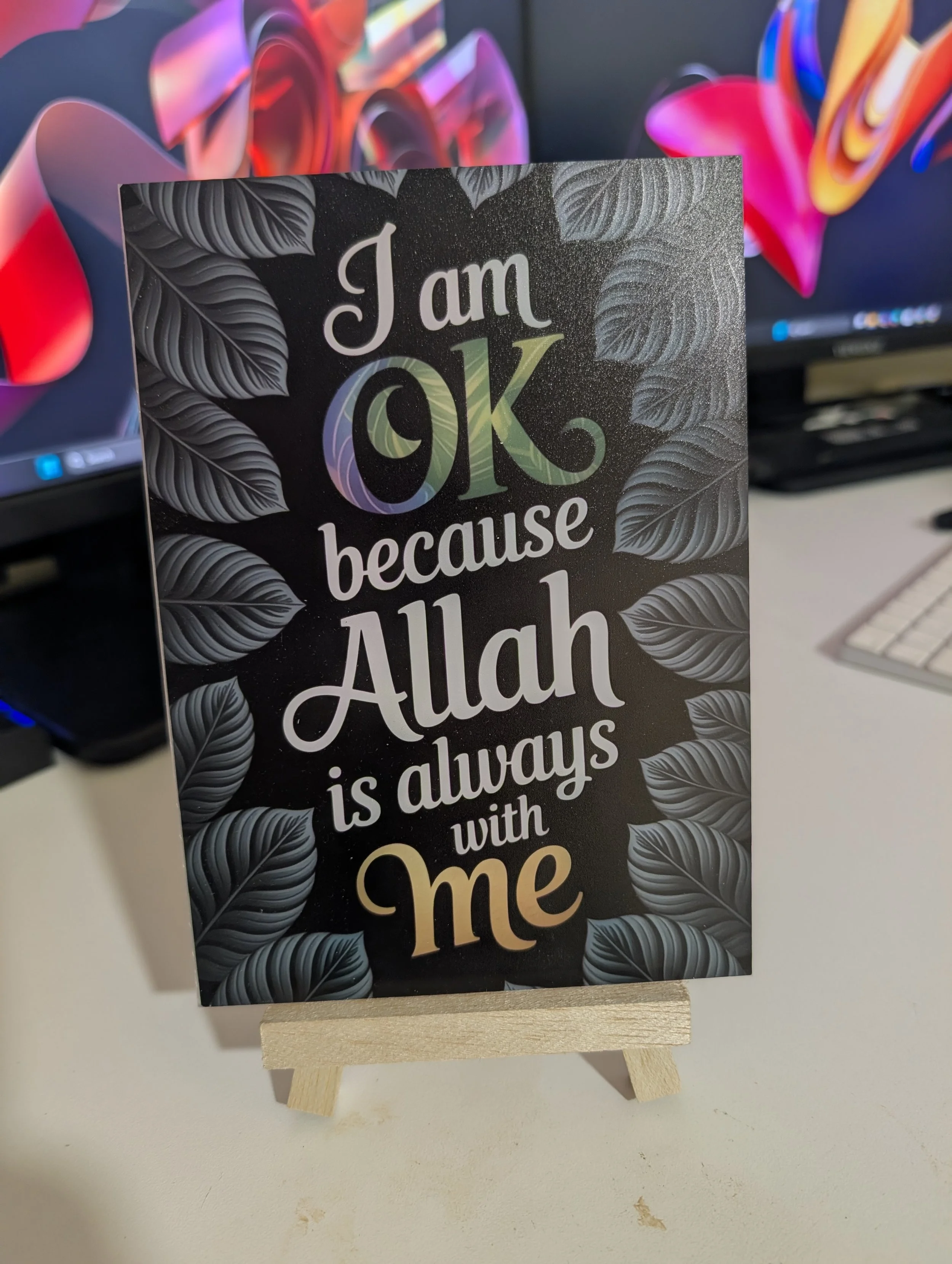 A decorative sign with the text 'I am OK because Allah is always with me', surrounded by leaf illustrations, placed on a small wooden stand on a white table, with computer monitors and colorful shapes in the background.