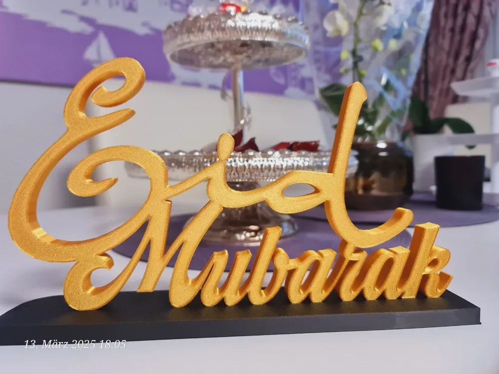 Decorative gold-colored Ramadan and Eid Mubarak greeting sign on a black base, with a blurred background of a dish stand and floral decor.