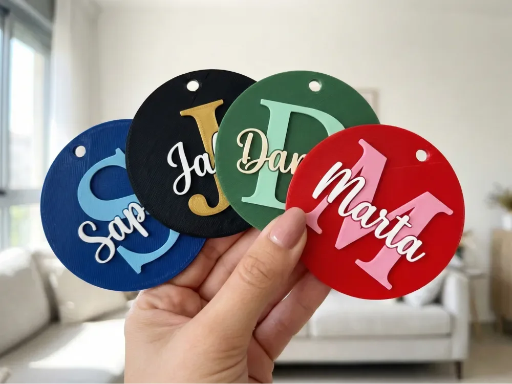 Four colorful round tags with letters and names, held in a person's hand. Each tag displays a name in cursive with a large initial letter. The names are Sap, Jack, Dan, and Martha, in blue, black, green, and red tags, respectively.
