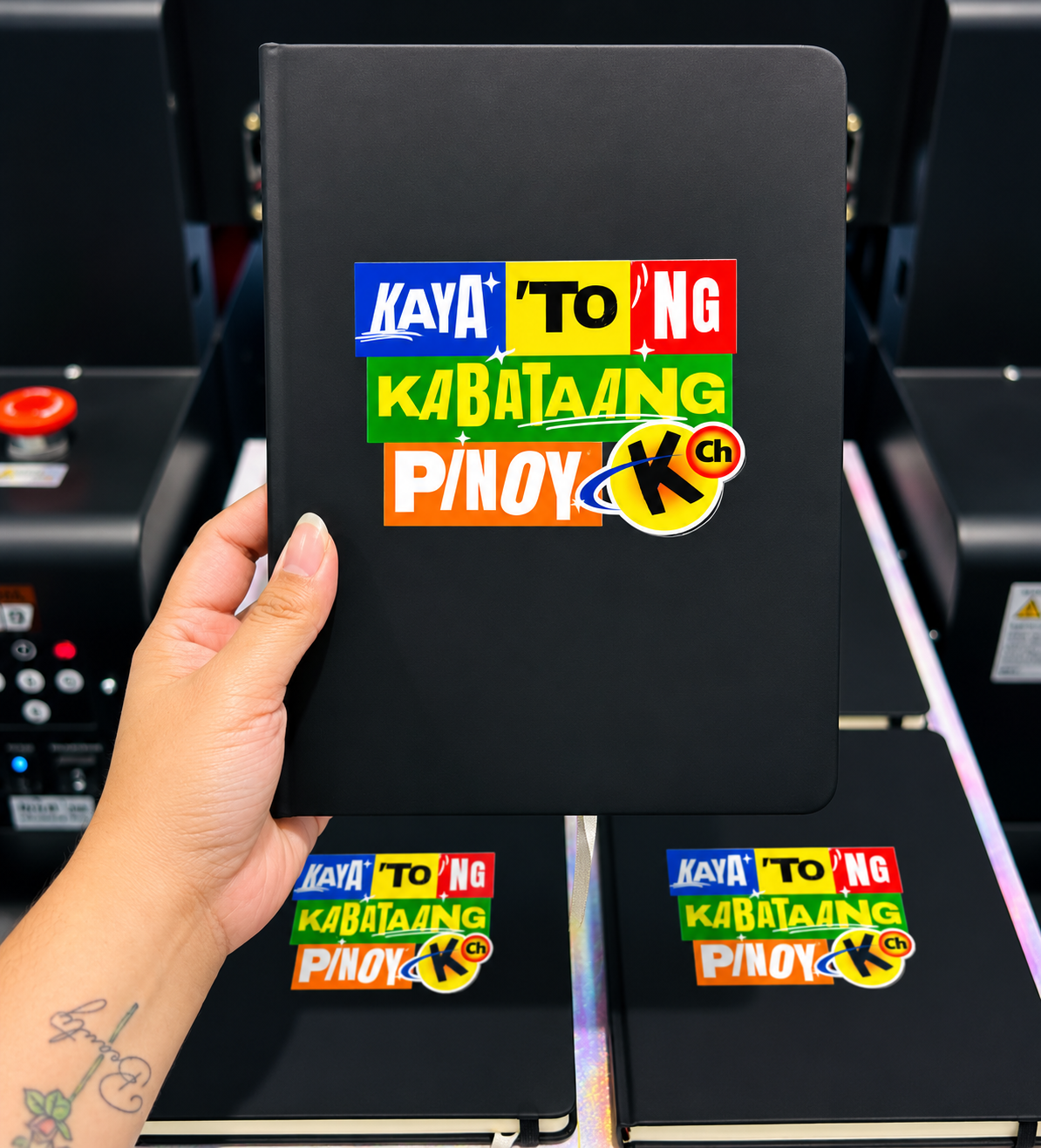 Hand holding a black notebook with colorful sticker designs that say 'KAYA 'TO' NG KABATAANG PINOY' and a small 'K Ch' logo, in front of other similar notebooks.