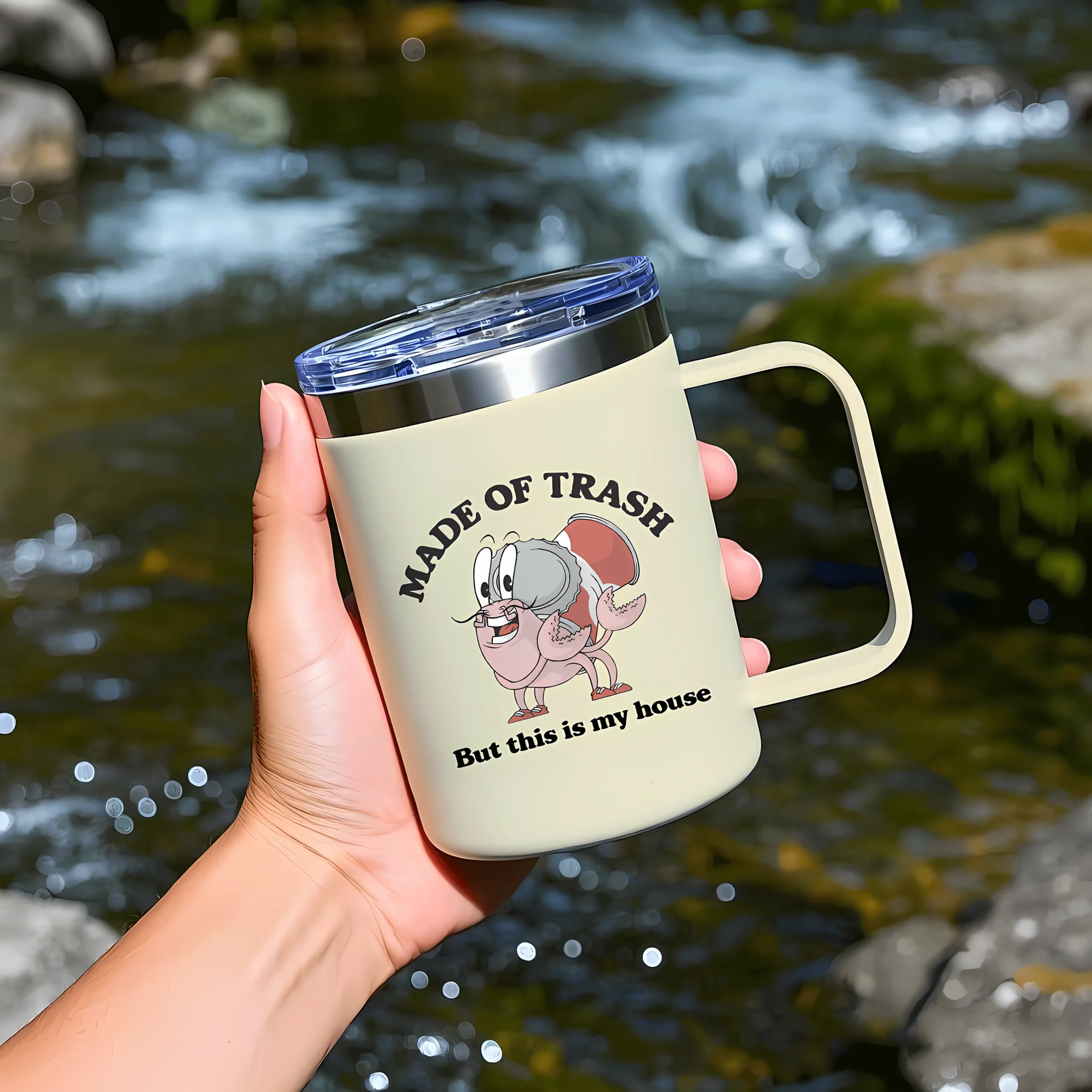 A person holding a travel mug with a cartoon of a brain with a beard and eyebrows, and the words "Made of Trash" and "but this is my house". The background shows a flowing stream with rocks.