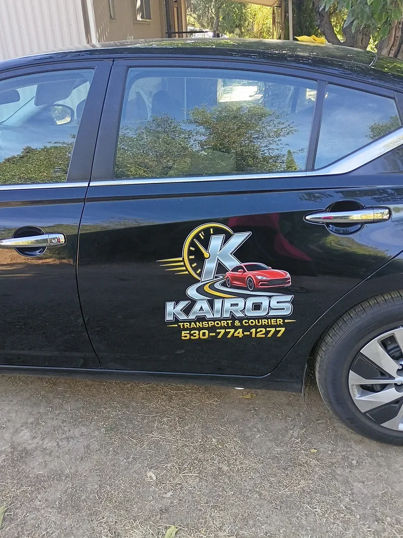 A black car with a logo on the door for Kairos Transport & Courier, including a red sports car graphic, a clock, and contact number 530-774-1277.