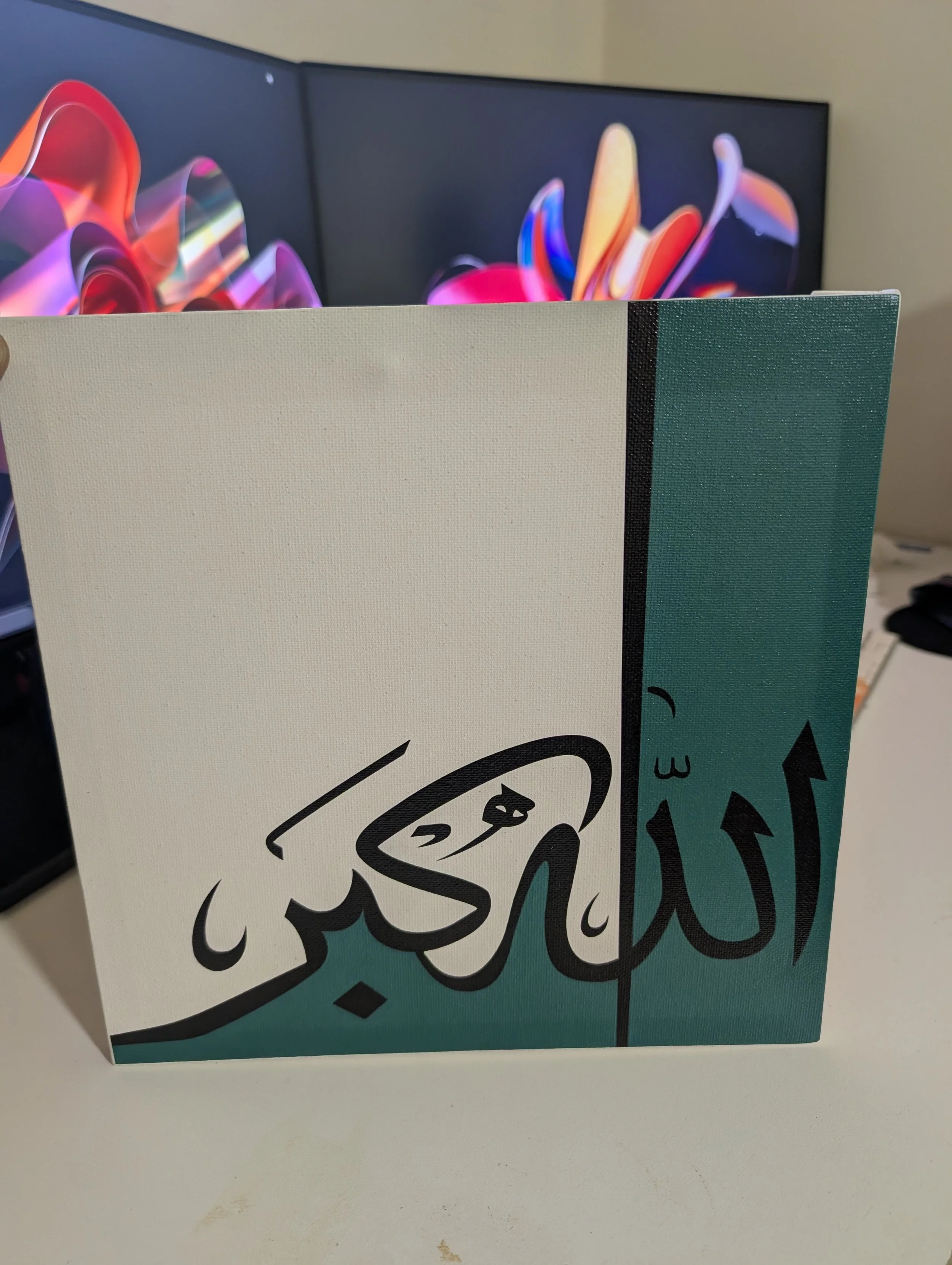 Close-up of a cardboard box with a colorful design featuring stylized flowers and leaves. The box is placed on a white surface with a computer monitor displaying a colorful abstract background in the background.