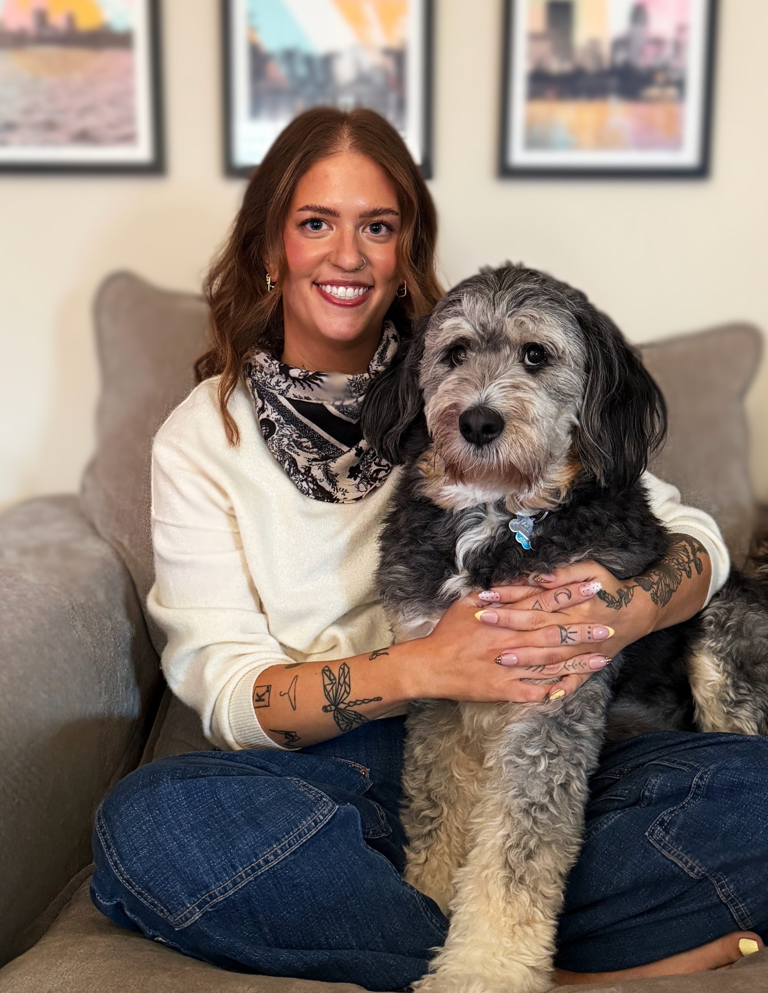 A woman with brown hair, tattoos on her arms and hands, and earrings, sitting on a couch holding a large, fluffy black and gray dog. The woman is smiling and looking at the camera, with a blurred background of framed pictures on a beige wall.