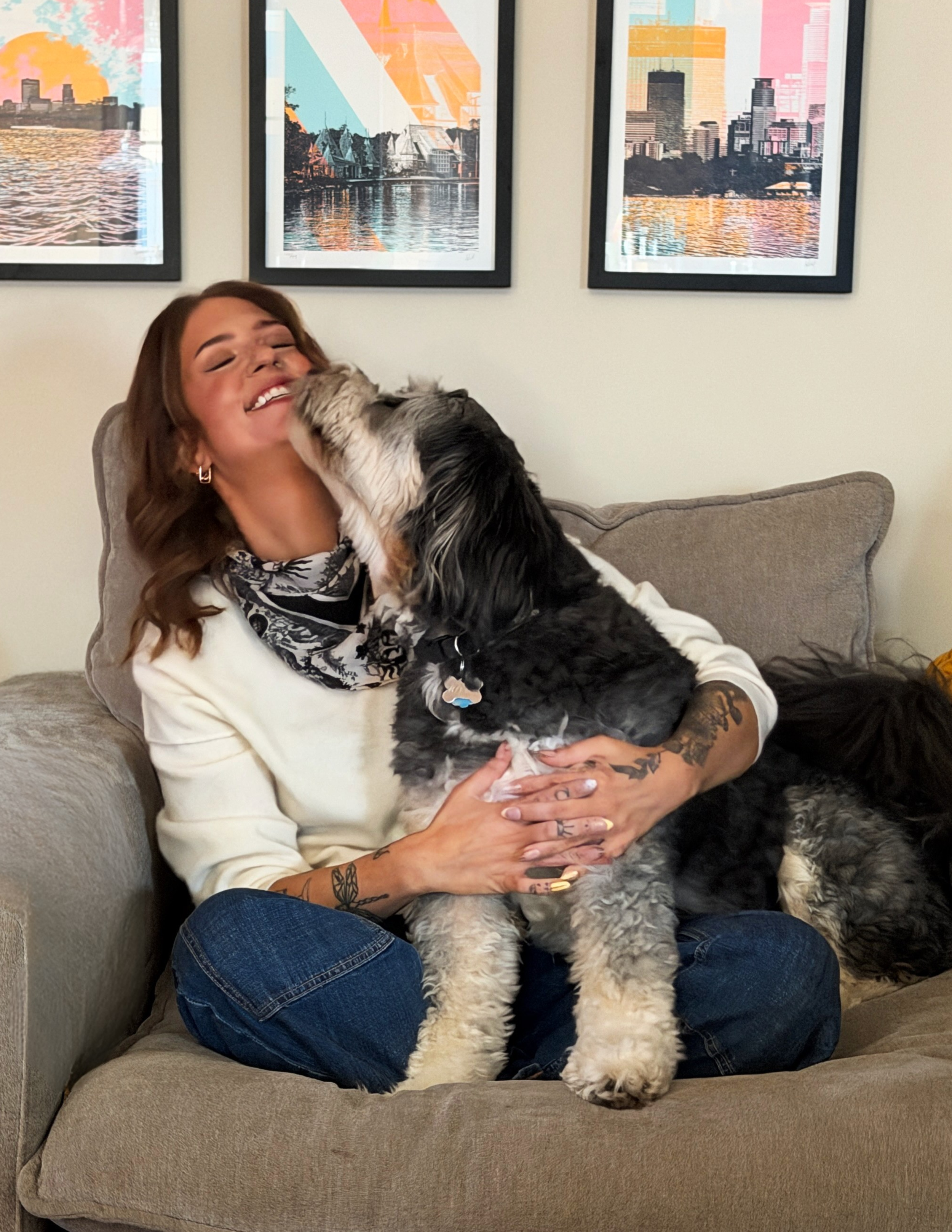 A woman sitting on a gray couch, smiling as a large black and white dog licks her face. The woman has tattoos on her arms and is wearing a cream-colored sweater and a patterned scarf. The background features three framed cityscape artworks on a beige wall.