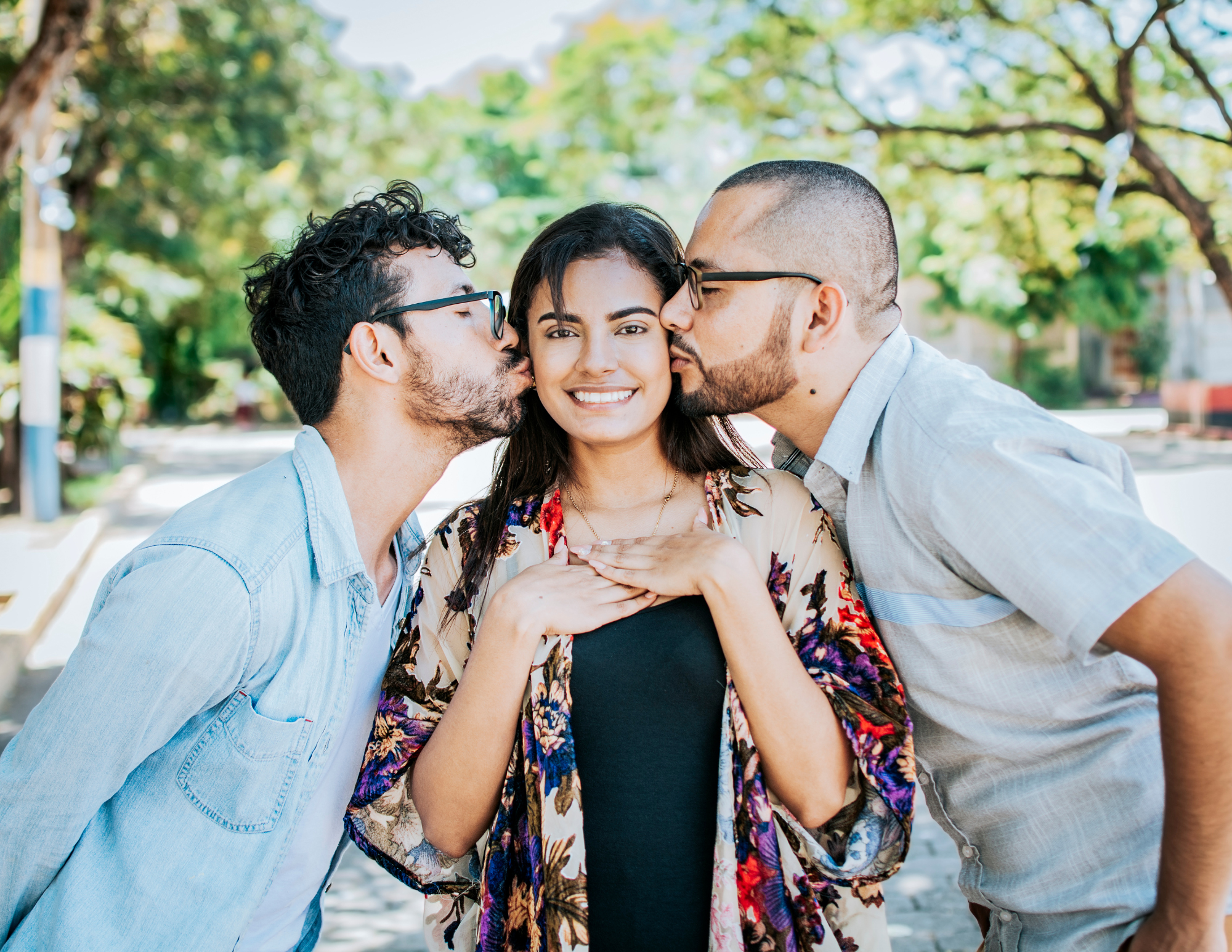A woman with dark brown hair stands smiling at the camera while 2 men stand on either side of her, kissing her cheeks