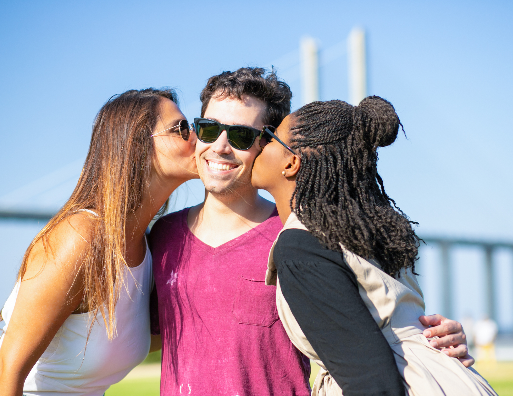 A man in sunglasses stands smiling and facing the camera while 2 women stand on either side of him, kissing his cheeks