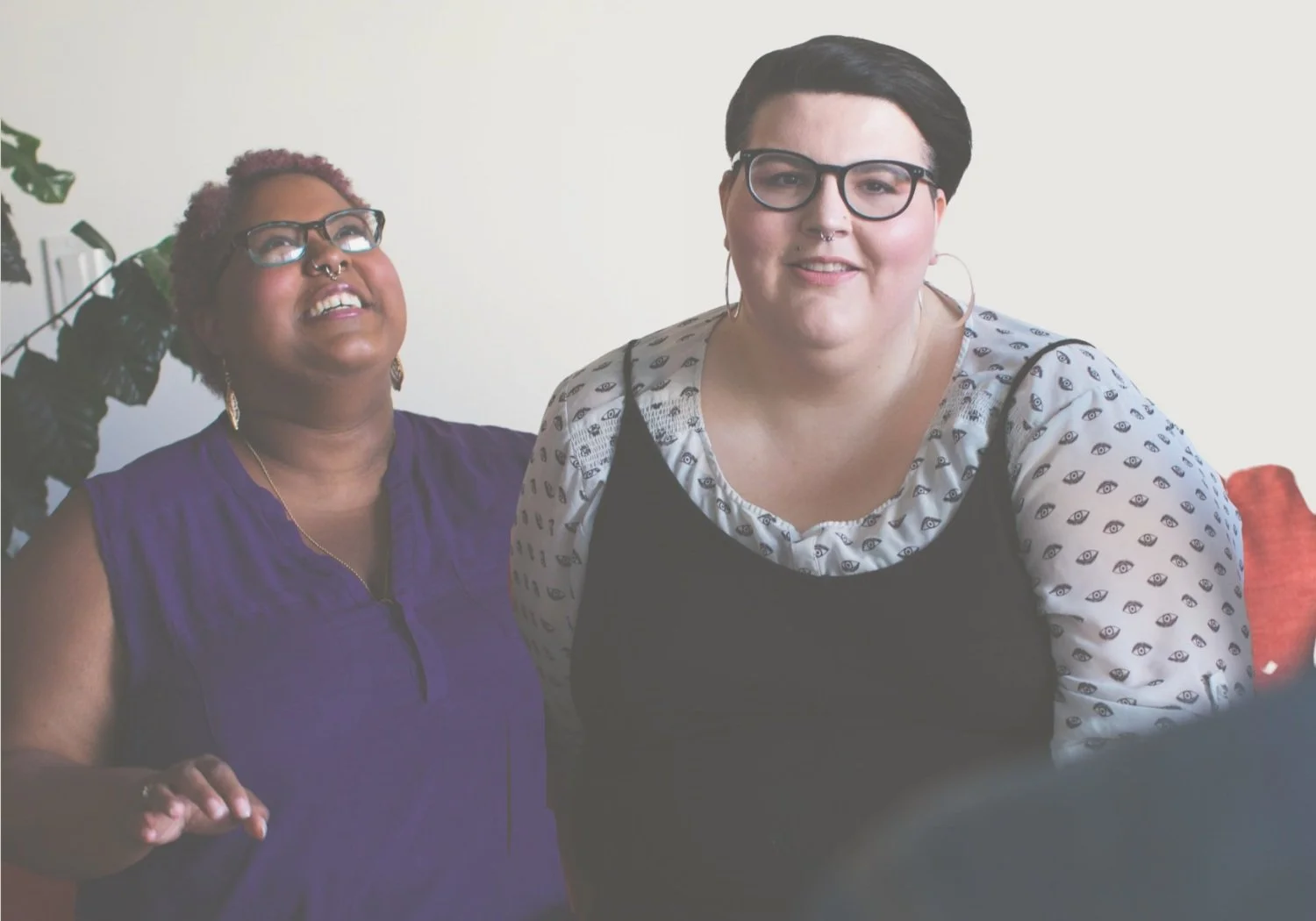 A queer, interracial, lesbian couple sit on a therapy couch, both smiling.