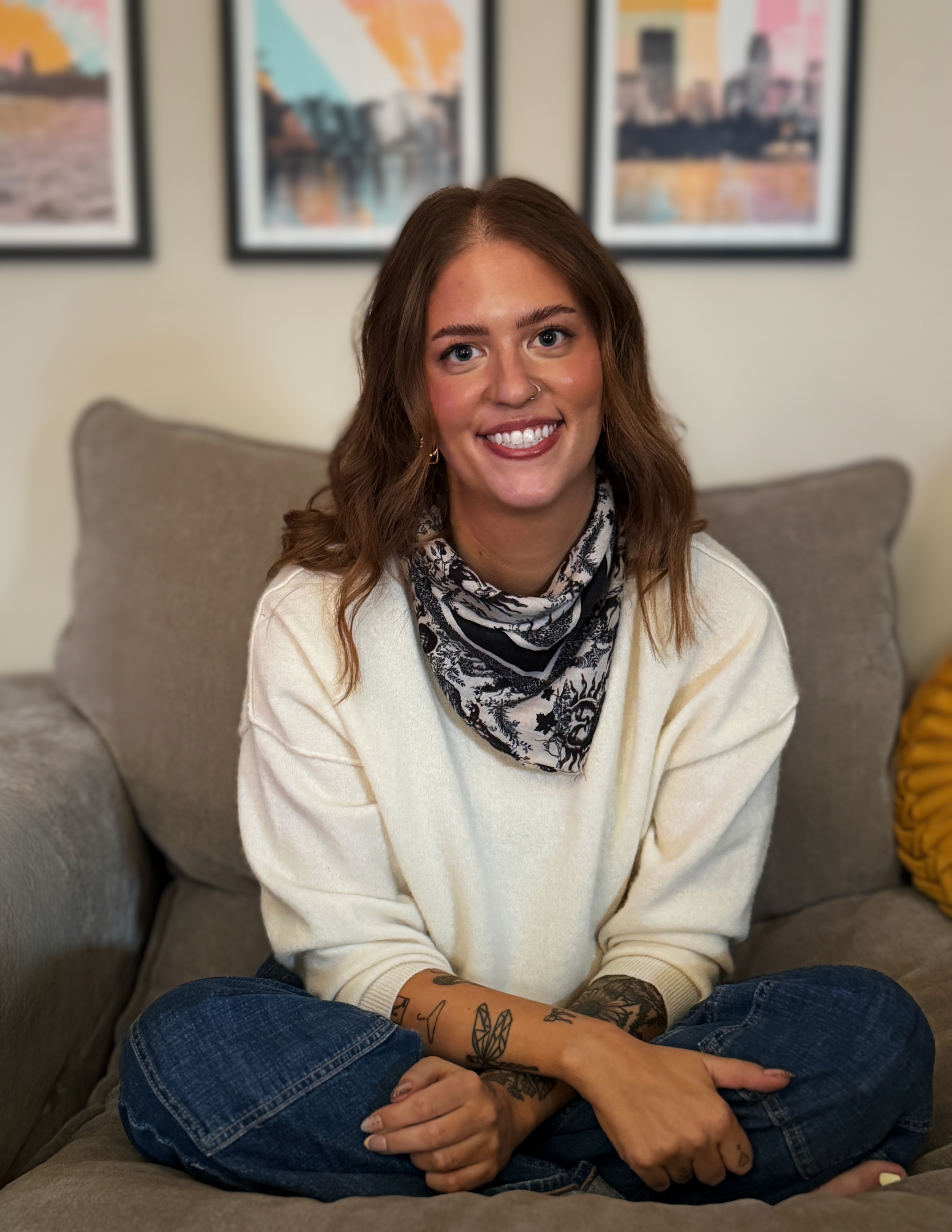 A woman with brown hair, blue eyes, and tattoos on her arms, sitting on a sofa with her hands clasped in her lap, smiling at the camera. She is wearing a cream-colored sweater and a black-and-white patterned scarf, with three framed photos of cityscapes in the background.