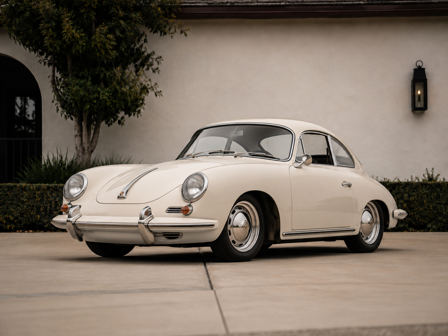 A vintage cream-colored Porsche 356 coupe parked on a driveway in front of a white wall with greenery and a black lantern.