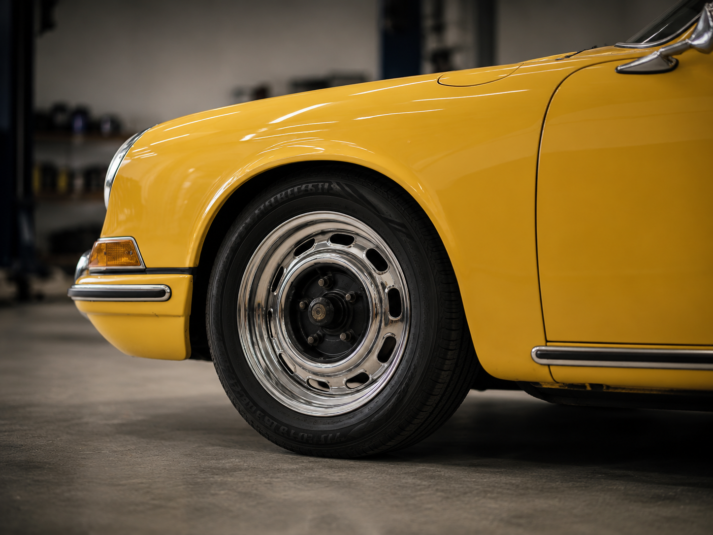 Close-up of a vintage yellow sports car's front section with chrome wheel and black tire in a garage.